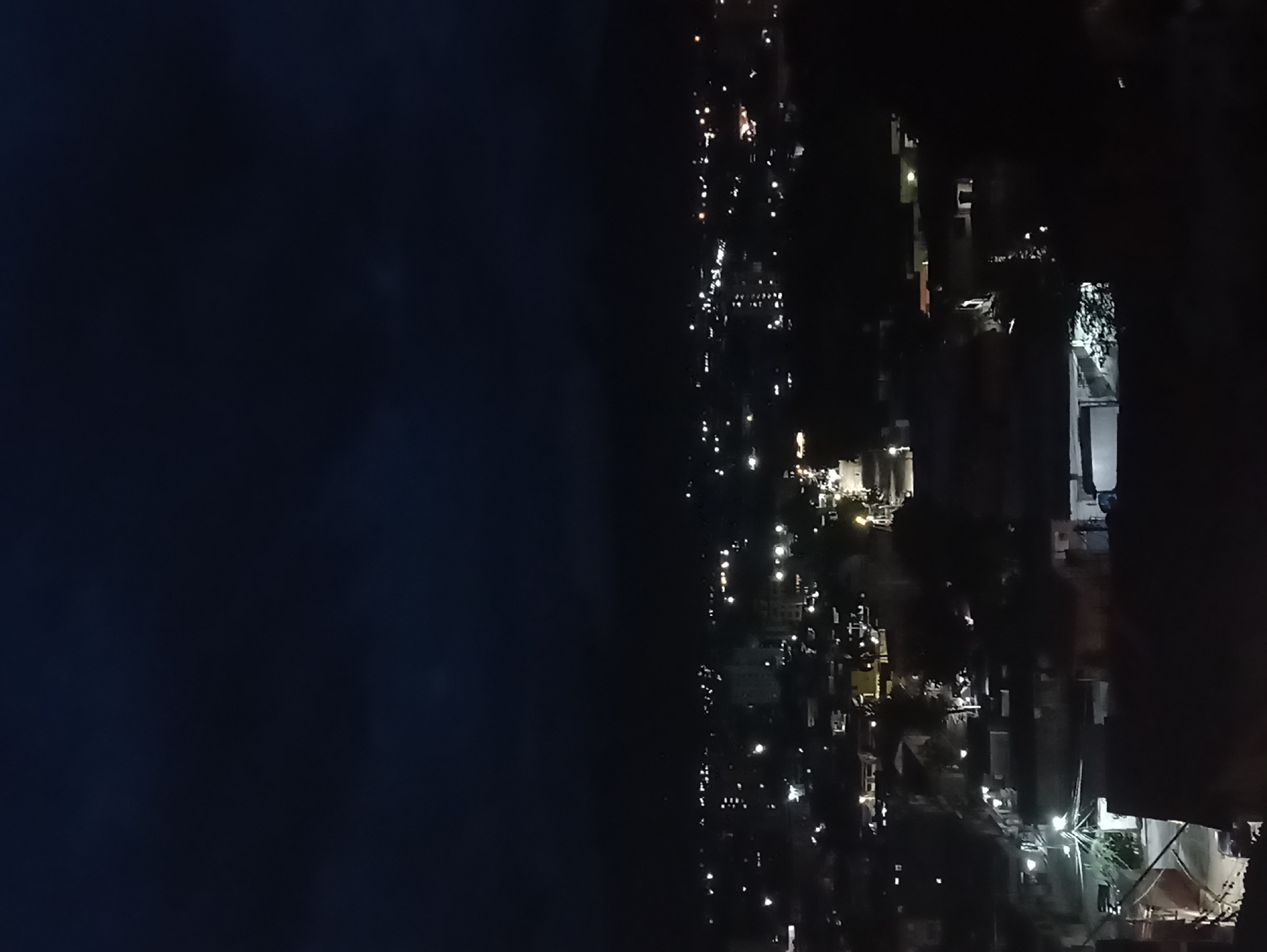 The image displays a high-angle, nighttime view of an urban or suburban area. The frame is distinctly divided by a sharp vertical line: the left side consists of a vast, uniformly dark expanse, while the right side is brightly illuminated by numerous artificial lights.
**Subjects and Objects:** The right side of the image is dotted with various artificial lights, which appear to originate from streetlights, buildings, and possibly vehicles. Faint outlines of structures and some indistinct vegetation are visible amidst the glow. No people are discernible in the scene.
**Activity or Event:** No specific activity or event is taking place; the image captures a static, serene moment of a settlement at night.
**Notable Details:**
* **Time of Day:** The pervasive darkness of the left side and the complete reliance on artificial illumination clearly indicate that the scene takes place at night.
* **Weather:** The dark expanse appears clear and calm, with no visible atmospheric disturbances like clouds or fog.
* **Location Cues:** The stark contrast between the dark, undefined area and the brightly lit urban section suggests either a view across a large body of water (such as a lake or sea) towards a city, or an overlook of a city against a dark, undeveloped landmass or the night sky. Given the context of "La Victoria, Venezuela," which is an inland city near Lake Valencia, the dark expanse could plausibly represent Lake Valencia at night, or simply the dark sky above the city. The density and arrangement of lights confirm a populated urban environment.
* **Emotions:** The scene evokes a sense of tranquility and the enduring presence of human activity amidst the night's vastness.
**Visible Text:** There is no discernable text within the image.