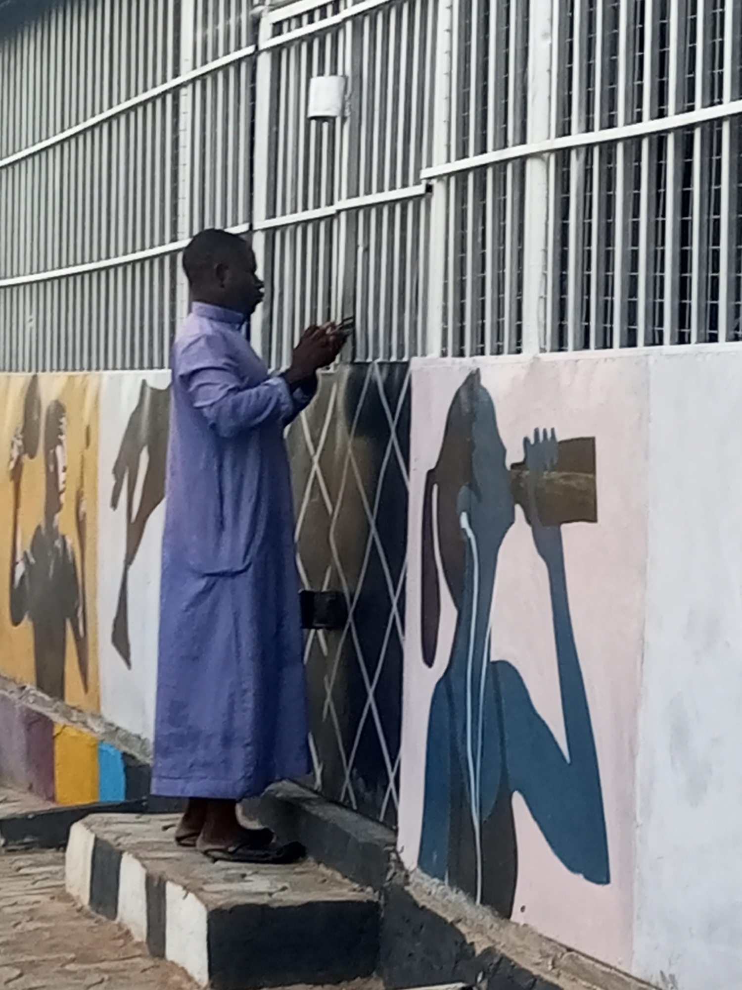 The image shows a man standing on a curb next to a wall with various artwork on it. The man is dark-skinned and is wearing a long, light blue robe and sandals. He is holding something in his hands, possibly a phone, and appears to be looking at it. The wall behind him is painted with several different designs, including silhouetted figures and geometric shapes. The setting appears to be an urban area with a modern building behind the artwork on the wall. The specific city and location are unknown.