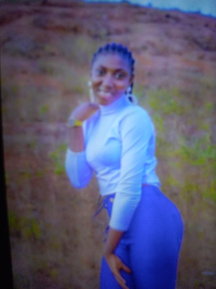 A young woman with dark hair in braids smiles toward the camera. She wears a white turtleneck and blue pants. Her left hand is raised to her cheek, and her right hand rests on her hip. The background appears to be a grassy hill or field, slightly blurred. The image has a soft focus, potentially due to the camera or lighting conditions.