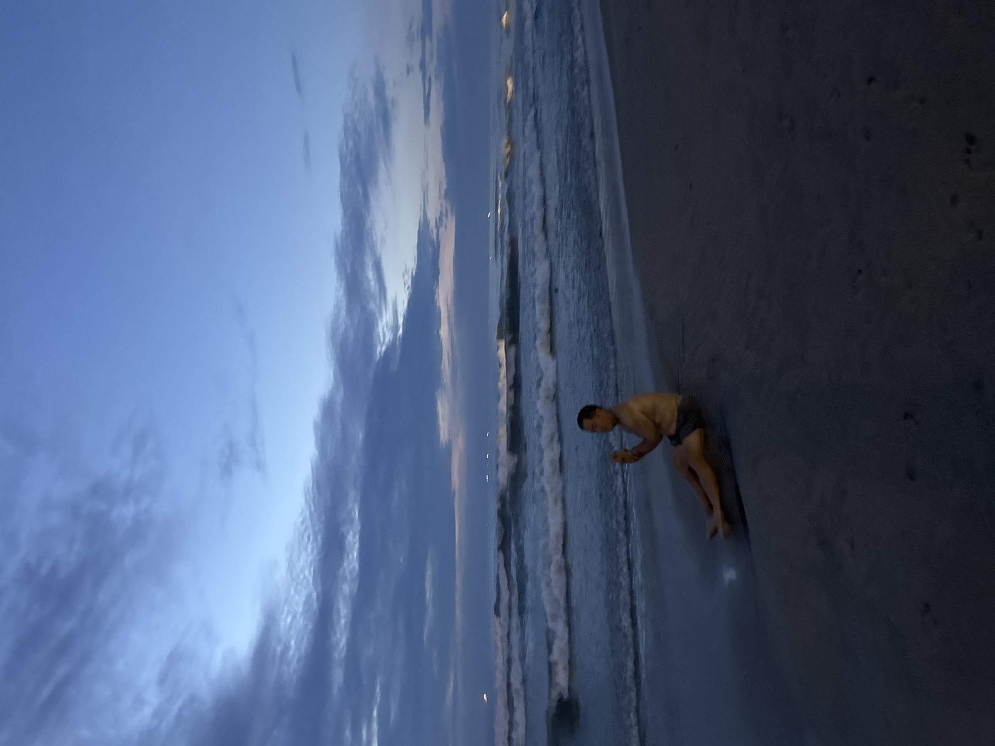 The image depicts a beach scene at dusk or dawn. A man is standing at the water's edge, bent over with his hands near the surface of the water. He is wearing dark swimwear. The beach sand appears dark, and the water is light in color, with visible waves. The sky is a mix of blue and cloudy. The location is likely somewhere in Đà Nẵng, Vietnam.