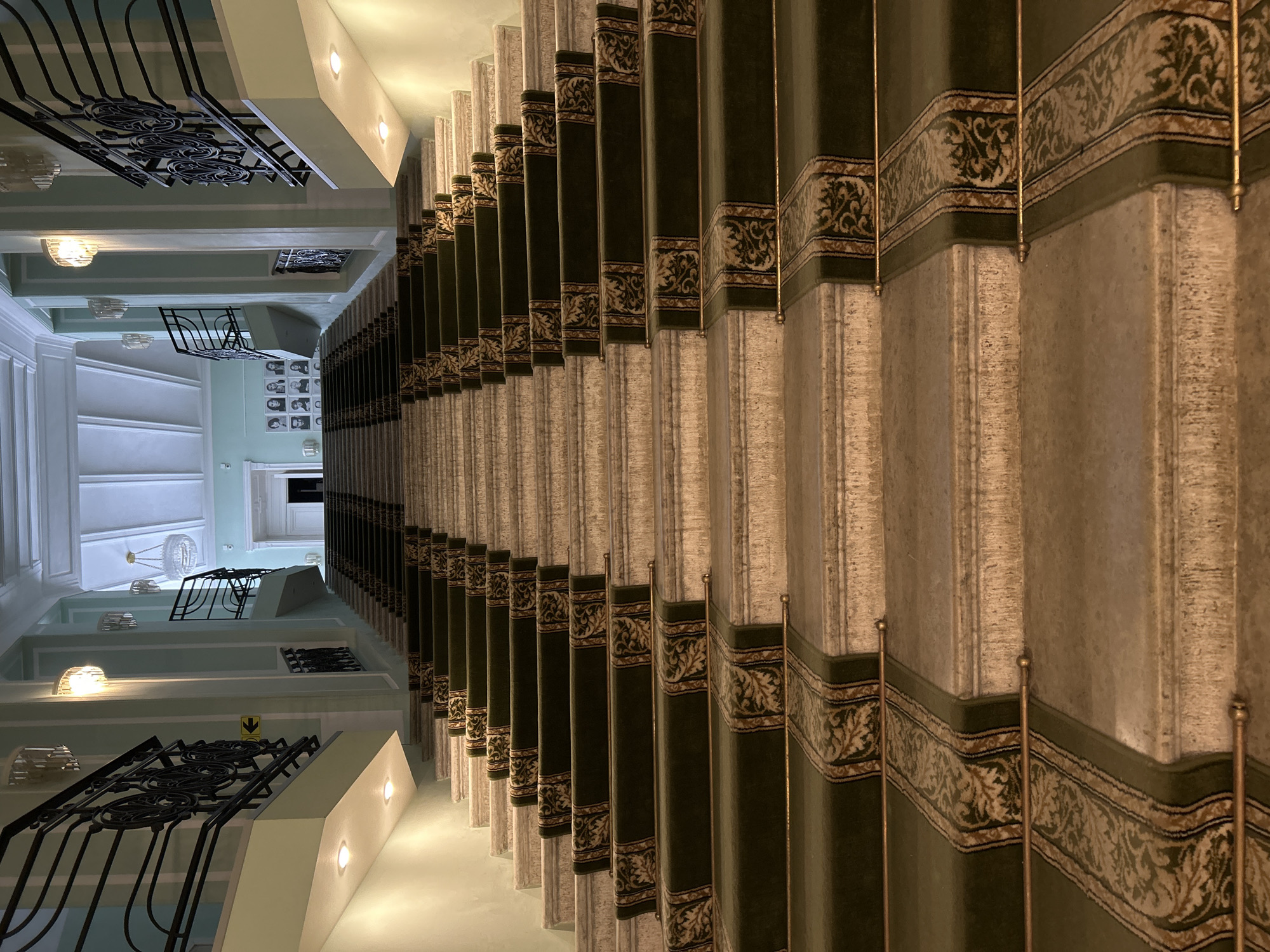 The image depicts a grand staircase in what is likely an upscale building. The steps are covered with a patterned carpet, featuring a green and beige color scheme. The walls are a pale green color, with decorative architectural details. Black wrought-iron railings are visible along the sides of the staircase. The lighting is a combination of natural light from above and artificial light fixtures. The style and architecture suggest the location is the Eliseyev Emporium, a historic building in Saint Petersburg, Russia, known for its ornate interior. No people are visible in the image.
