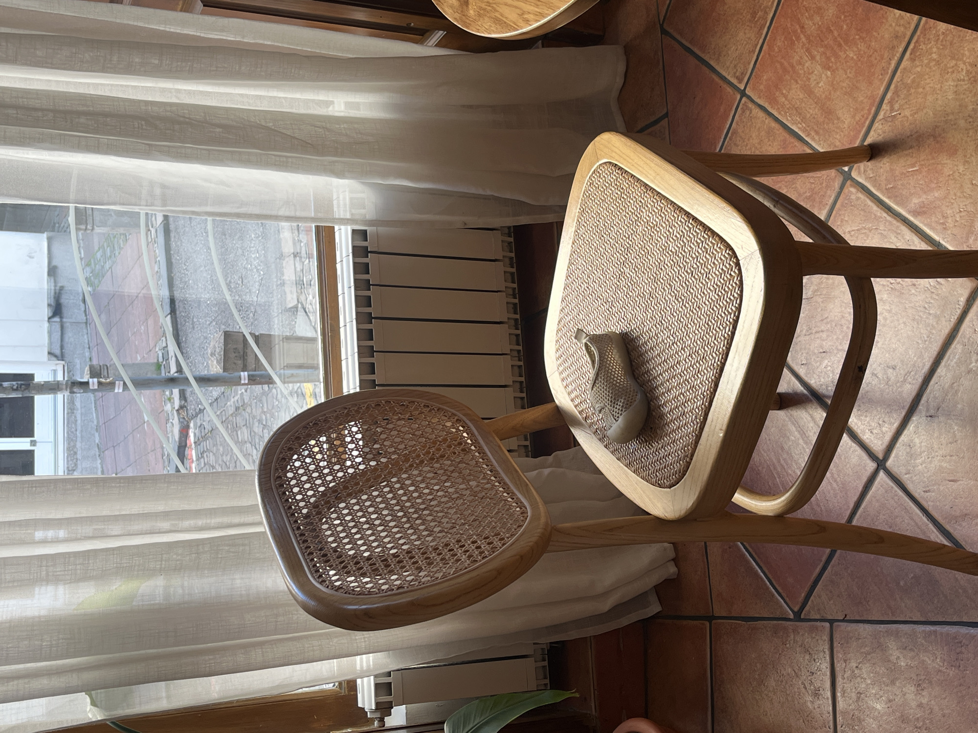 A light brown wooden chair with a woven cane back and seat is positioned in front of a window with white curtains. A small, light-colored shoe rests on the seat of the chair. The floor is tiled with reddish-brown square tiles. A radiator is visible beneath the window. Outside the window, a street and building are visible, suggesting an urban setting. The scene appears to be indoors, possibly in a cafe or a residential space. The lighting suggests it is daytime. There is no indication that this scene is located in Belgrade, Serbia.