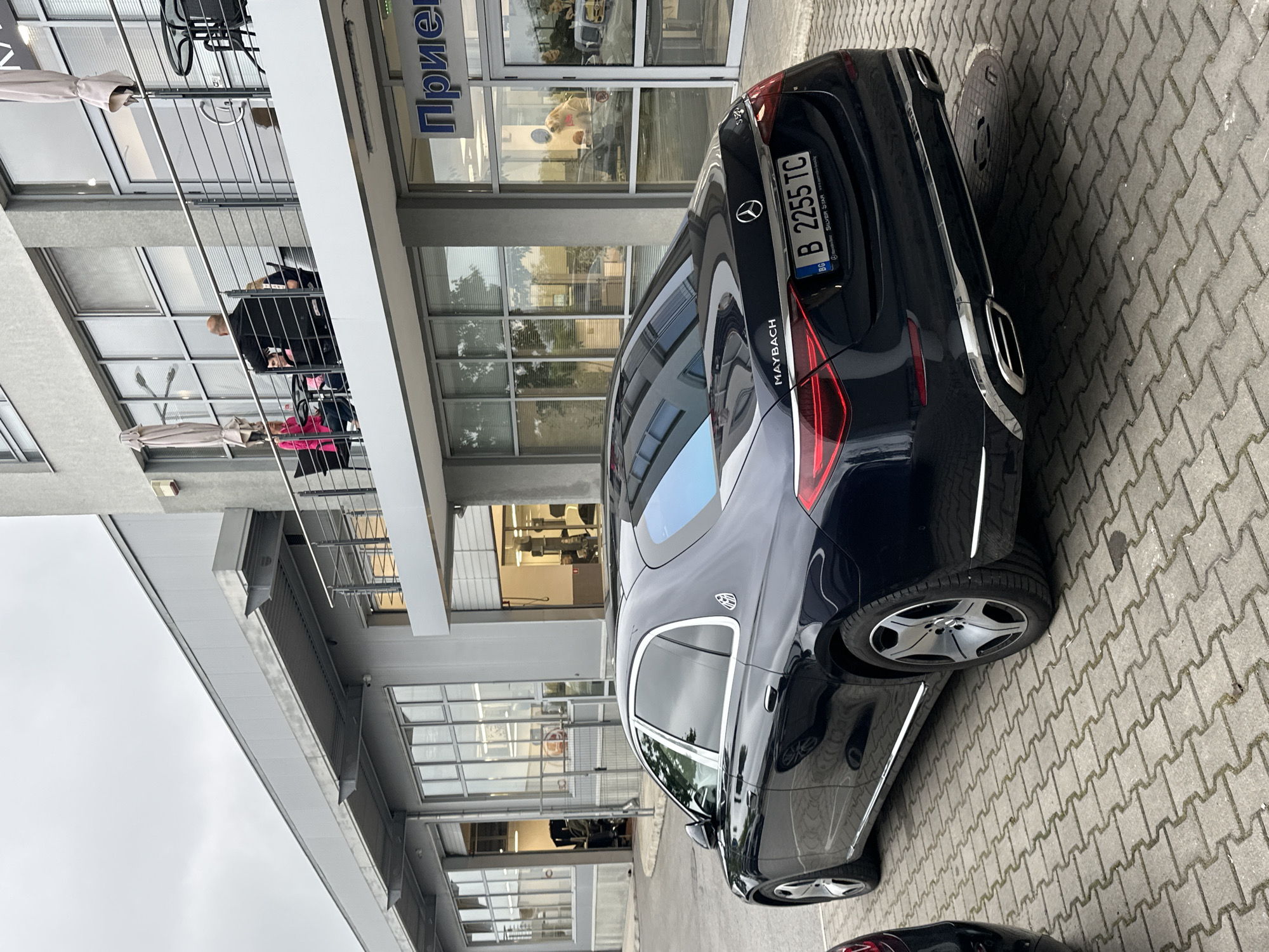 The image depicts a dark-colored Mercedes-Benz Maybach sedan parked on a paved surface in front of a modern commercial building. The ground is covered with grey interlocking pavers, and a circular manhole cover is visible near the car's rear.
The luxury sedan is prominently featured, displaying the "MAYBACH" emblem on its trunk lid. It bears a Bulgarian license plate, "B 2255 TC", and the license plate frame includes text such as "Mercedes-Benz," "Super Star," and "www.superstar.bg," indicating a dealership. The car's reflections show parts of the surrounding building.
The multi-story building features a contemporary design with extensive glass windows, doors, and metal railings on balconies or walkways. On an upper balcony, several people are visible; one individual in a black jacket sits at a table with their back to the viewer, while another in a bright pink top is partially seen. Two beige parasols are also present on this balcony, suggesting an outdoor seating area. A sign on the building's facade displays partially visible Cyrillic text, "Прие", which likely is part of "Прием" (Reception).
The scene is set during the daytime under an overcast sky, providing soft, diffused lighting. The overall environment suggests an urban or commercial area, possibly a car dealership or a business complex in Bulgaria, based on the license plate and visible text.