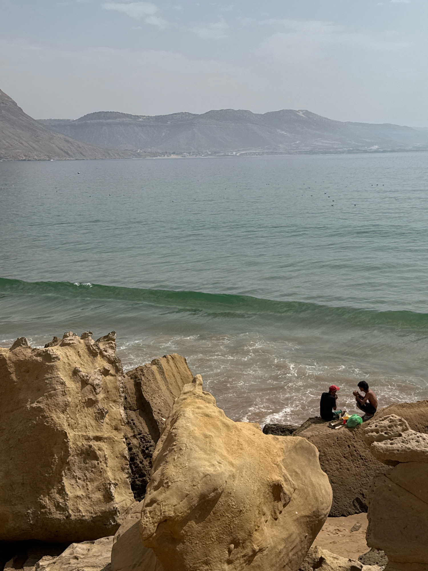 Two individuals are seated on a rocky outcrop overlooking the ocean in Imsouane, Morocco. The person on the left wears a red cap and a dark shirt, while the person on the right is shirtless. They appear to be interacting with objects on the ground between them, possibly food or supplies, and a green bag rests nearby. The rocky shore is composed of large, eroded sandstone formations. The ocean exhibits a gentle wave pattern with whitewater foam at the shore. In the distance, a hazy mountain range stretches across the horizon under a pale sky. Several dark specks, likely birds, are visible on the water's surface.