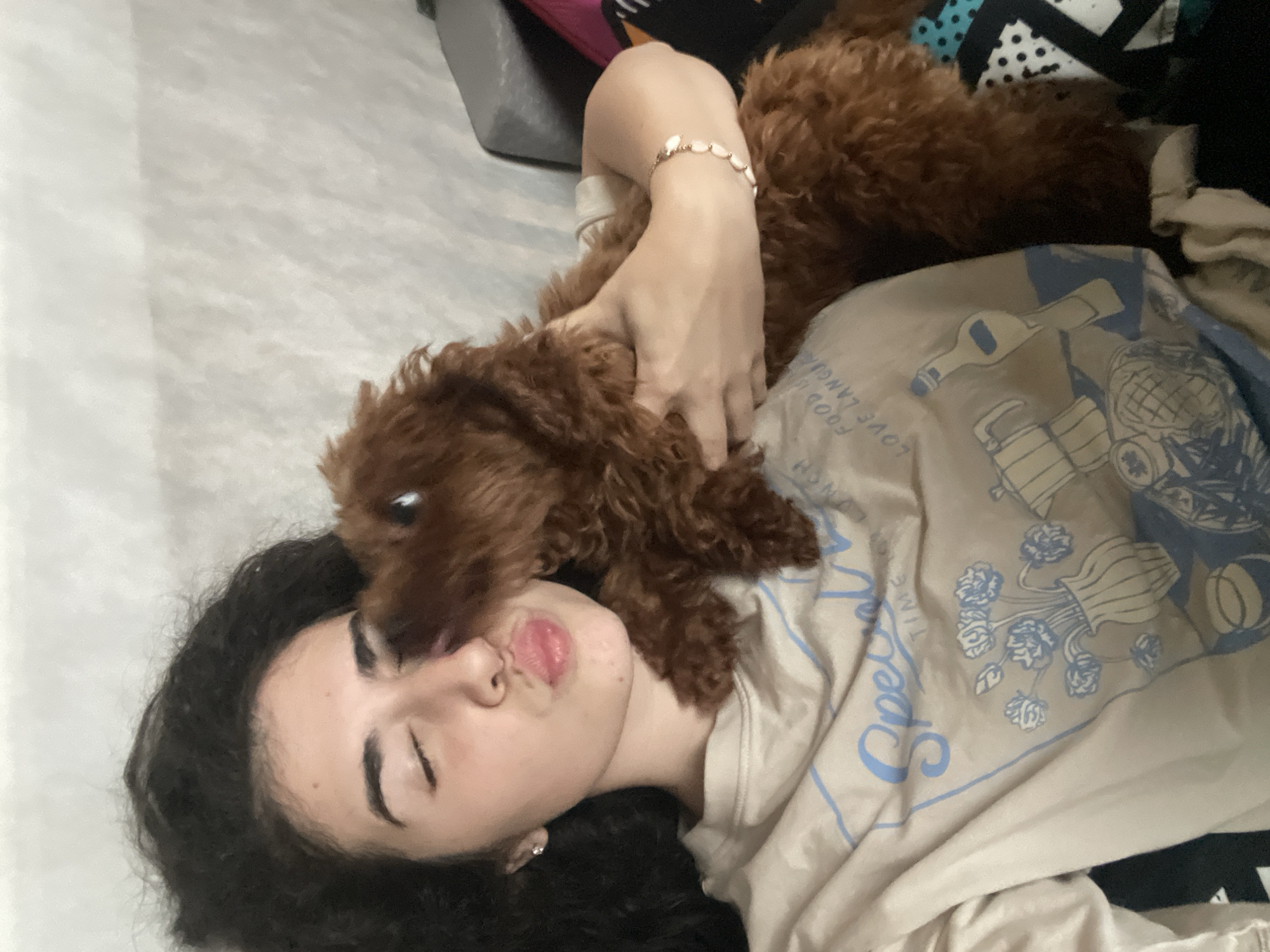A woman lies on a patterned surface indoors, with a brown poodle resting on her chest. The woman has dark curly hair and closed eyes, with her lips puckered. She wears a light-colored t-shirt with blue designs and text. Her left arm is bent, with her hand near the dog. She is wearing a bracelet. Part of a grey pillow is visible in the upper left corner. The location cannot be determined without further information.