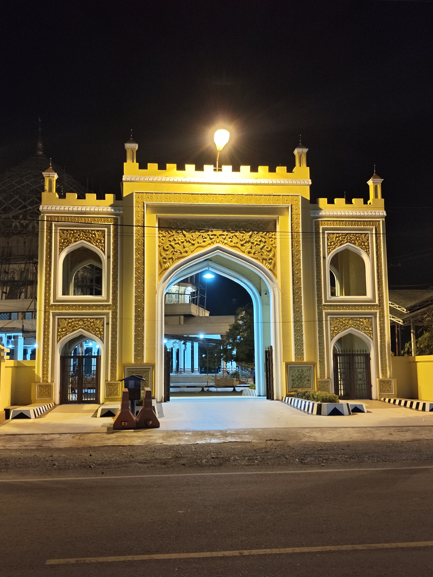 The image depicts the entrance gate of a mosque in Cotgadong, Indonesia, at night. The gate is elaborately decorated with yellow and gold ornamentation and features arched doorways. Two pedestrian entrances with metal gates are visible on either side of the main archway. In front of the gate, there are two brown traffic barriers. The surrounding area is dimly lit, with a street lamp illuminating the scene. The ground in front of the gate is paved, and a road with yellow lane markings is in the foreground. In the background, construction scaffolding is visible, suggesting ongoing development or renovation.