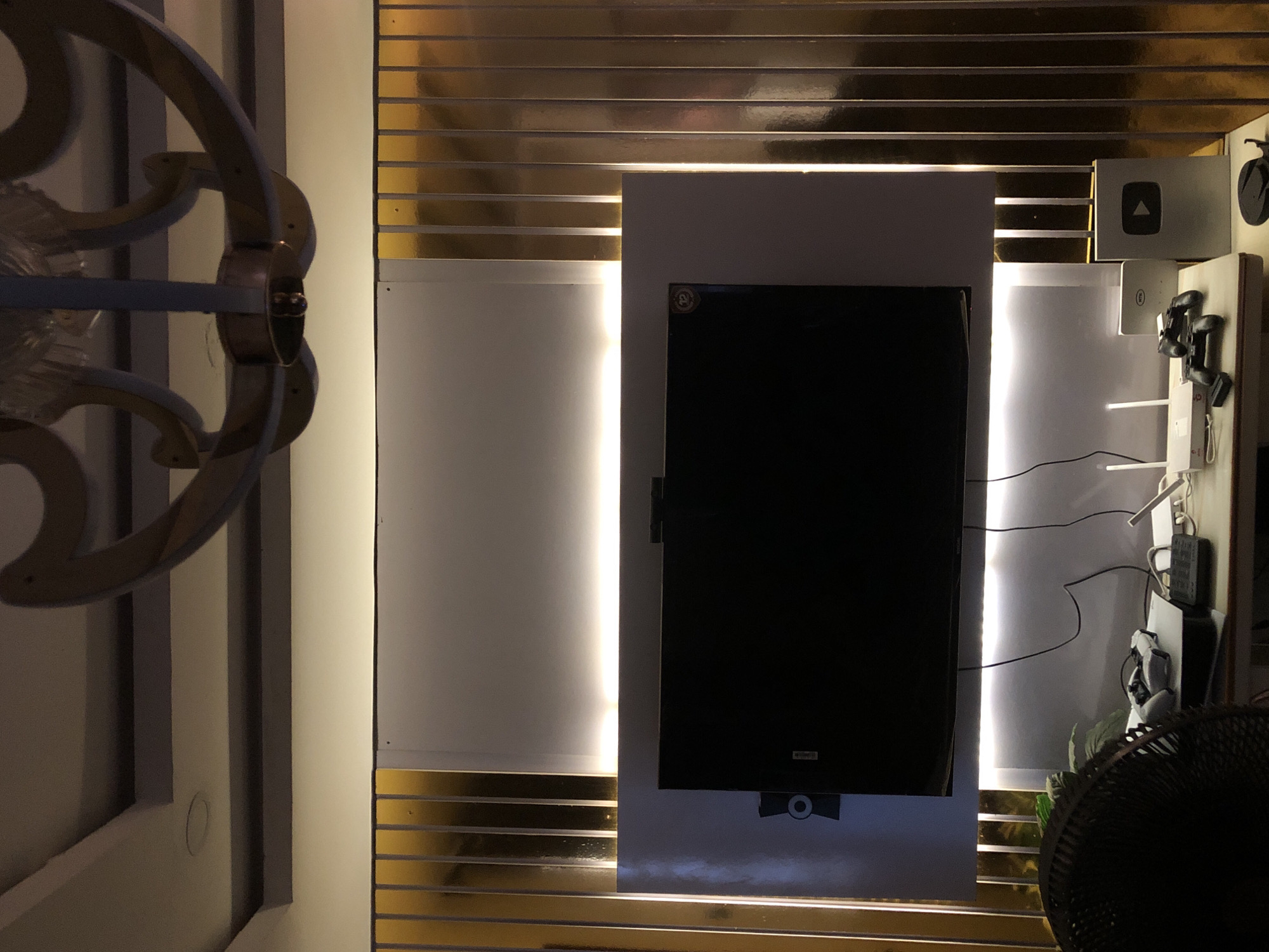 This indoor image presents a modern entertainment setup, likely in a dimly lit room or during evening hours, relying heavily on artificial illumination. A large, black-screened television is centrally mounted on a vertical white panel. Flanking the television are two prominent vertical light strips, emitting a bright, warm glow that illuminates the white panels.
Behind and surrounding the central white panel, the wall features horizontal, reflective gold-colored strips, creating a layered and somewhat opulent backdrop. On the far left, a large, ornate decorative element, possibly a wall sconce or part of a partition, with curved, metallic-toned structures, is partially visible.
To the right of the television, various electronic devices are organized on shelves or mounts. A notable detail is a silver plaque displaying the iconic YouTube Play Button logo (a triangle within a square), suggesting an award or achievement. Below this, multiple game controllers or similar devices are visible, along with a network of black cables. A black fan is partially visible in the bottom right corner of the frame.
The overall scene suggests a contemporary interior design focused on media consumption. Small, indistinct text or logos appear on the television screen itself (a faint white label at the bottom center and a circular logo on the upper left corner) and on some of the electronic devices, but are not legible. No people or specific activities are depicted.