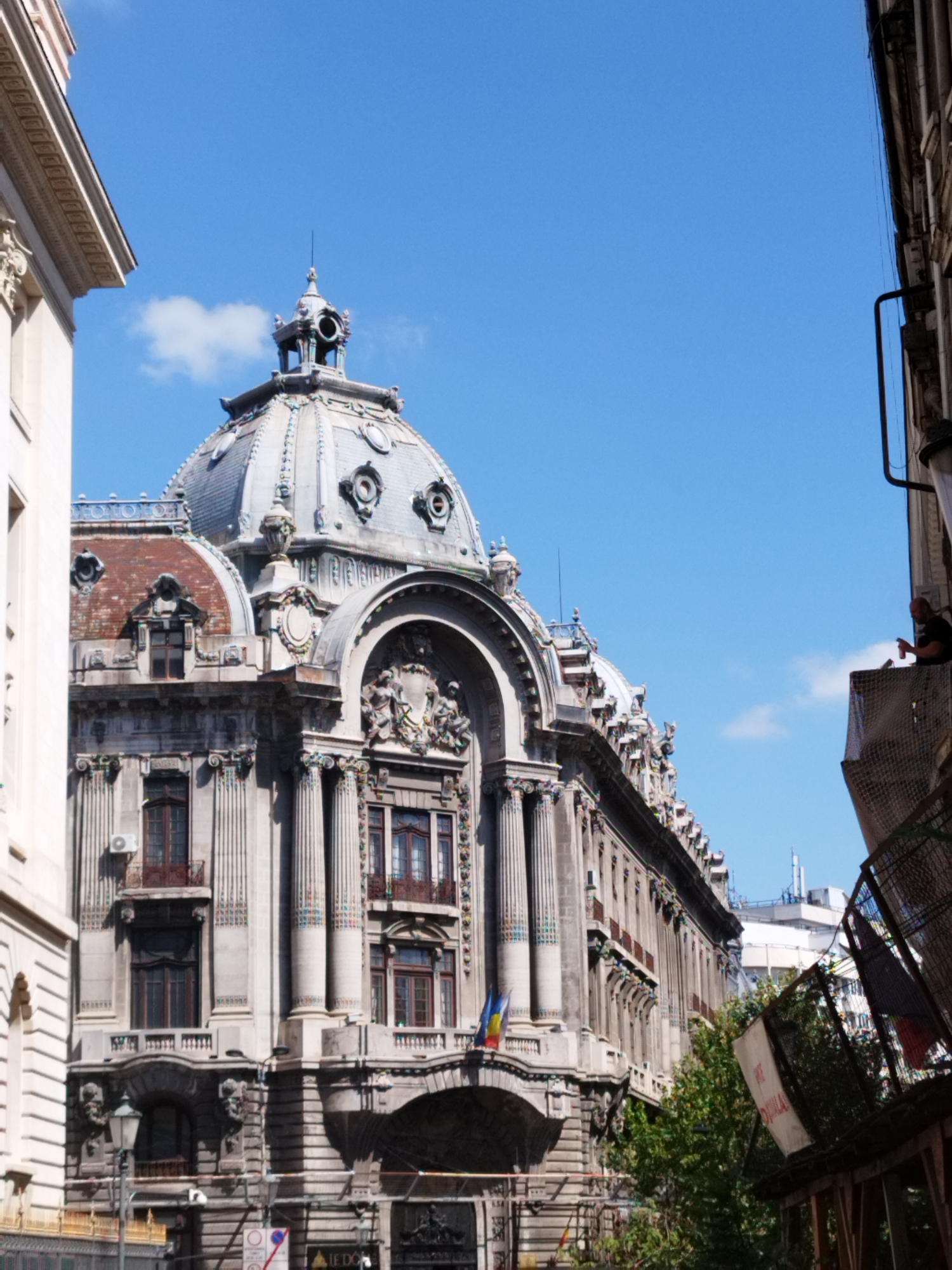 The image depicts the imposing facade of the CEC Palace in Bucharest, Romania, on a clear, sunny day with a bright blue sky and a few wispy clouds. The palace, an architectural masterpiece of Beaux-Arts style, dominates the frame with its intricate details and grand dome. The lower portion of the image shows a street scene, with hints of foliage and scaffolding or barriers in the foreground.
On the far right side of the image, a man is visible on a balcony or elevated platform, looking down and holding a mobile phone. He is wearing a dark t-shirt. The building exhibits a rich array of architectural elements, including ornate columns, arched windows, decorative sculptures, and a prominent dome topped with a small spire. Several windows are visible, some with balconies. Romanian flags are displayed on the building. The street level shows signs, a lamp post, and a security camera. The overall impression is one of historical grandeur meeting contemporary urban life.