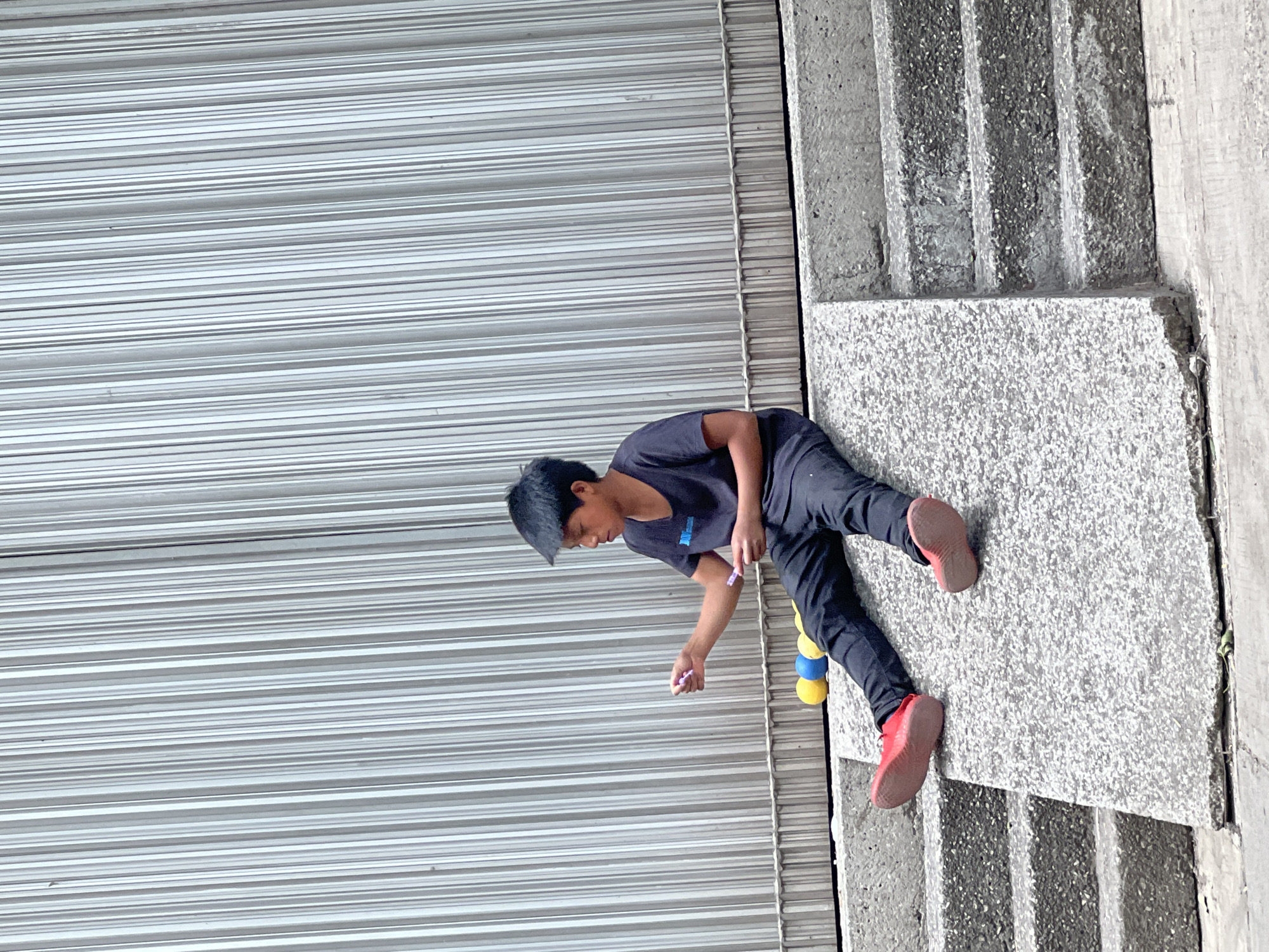A child in dark clothing and red shoes is on a gray concrete structure beside a corrugated metal wall. The setting appears to be an urban environment with the presence of concrete steps and a building facade, possibly in Mexico City, Mexico. The child seems to be interacting with something on the ground near the wall, possibly with small items in their hand. There is a yellow and blue cylindrical object nearby.