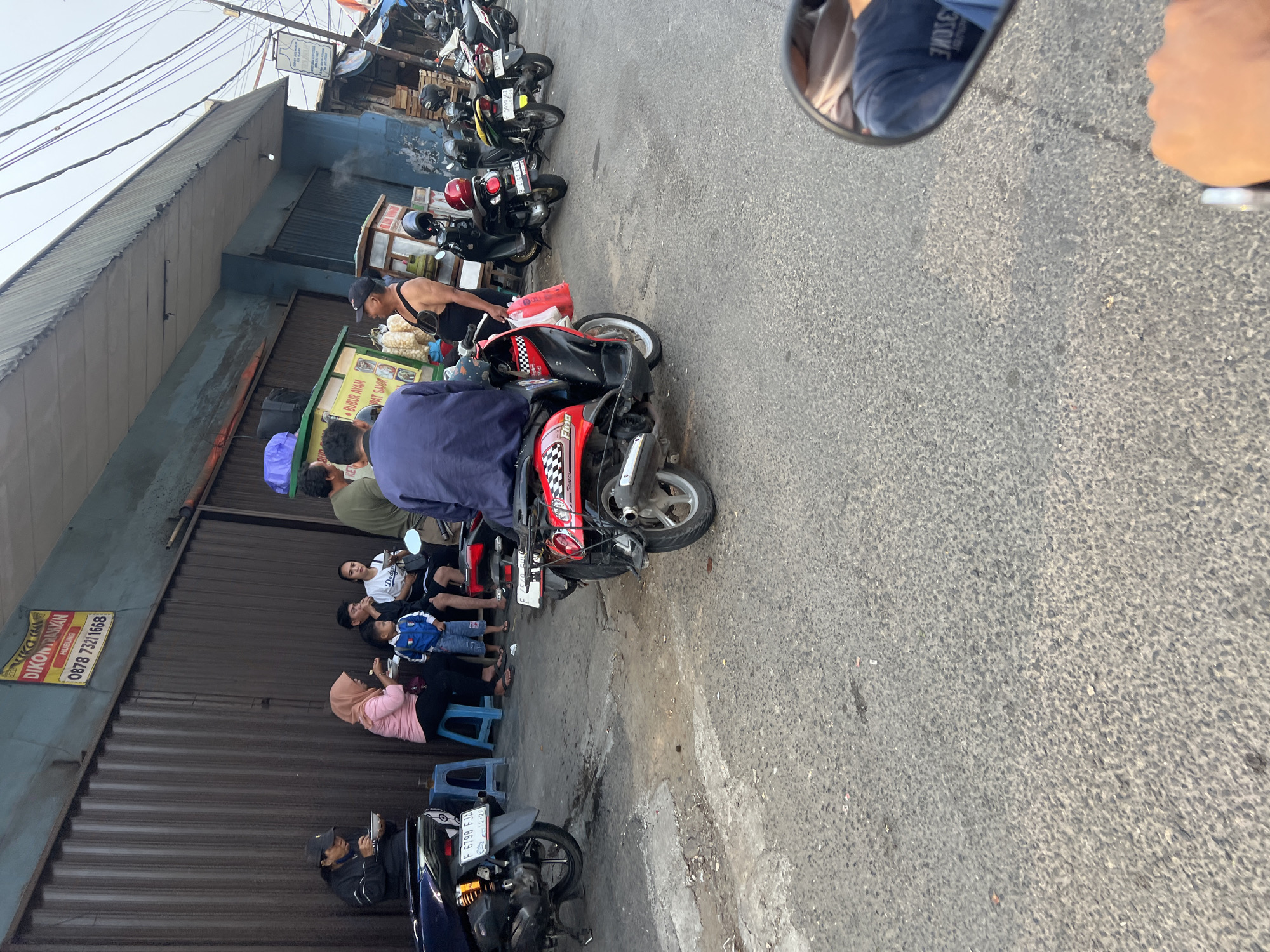 A group of people are gathered in front of a building with a corrugated metal facade in Citeureup, Indonesia. Several motorcycles are parked on the asphalt road. Two individuals are seated on blue plastic stools, eating from bowls. A man in a blue jacket is seated on a red and black scooter, while another man wearing a black tank top and shorts stands nearby holding a red plastic bag. A woman in a pink hijab and a pink shirt is also seated, eating. A young boy in a blue shirt and shorts is standing between the woman and another seated individual. The building has a blue painted section above the metal facade and a sign with red and yellow lettering is visible. Power lines are strung overhead, and wooden crates are stacked to the right of the building. The scene appears to be a casual gathering, possibly a street food stall or local eatery.