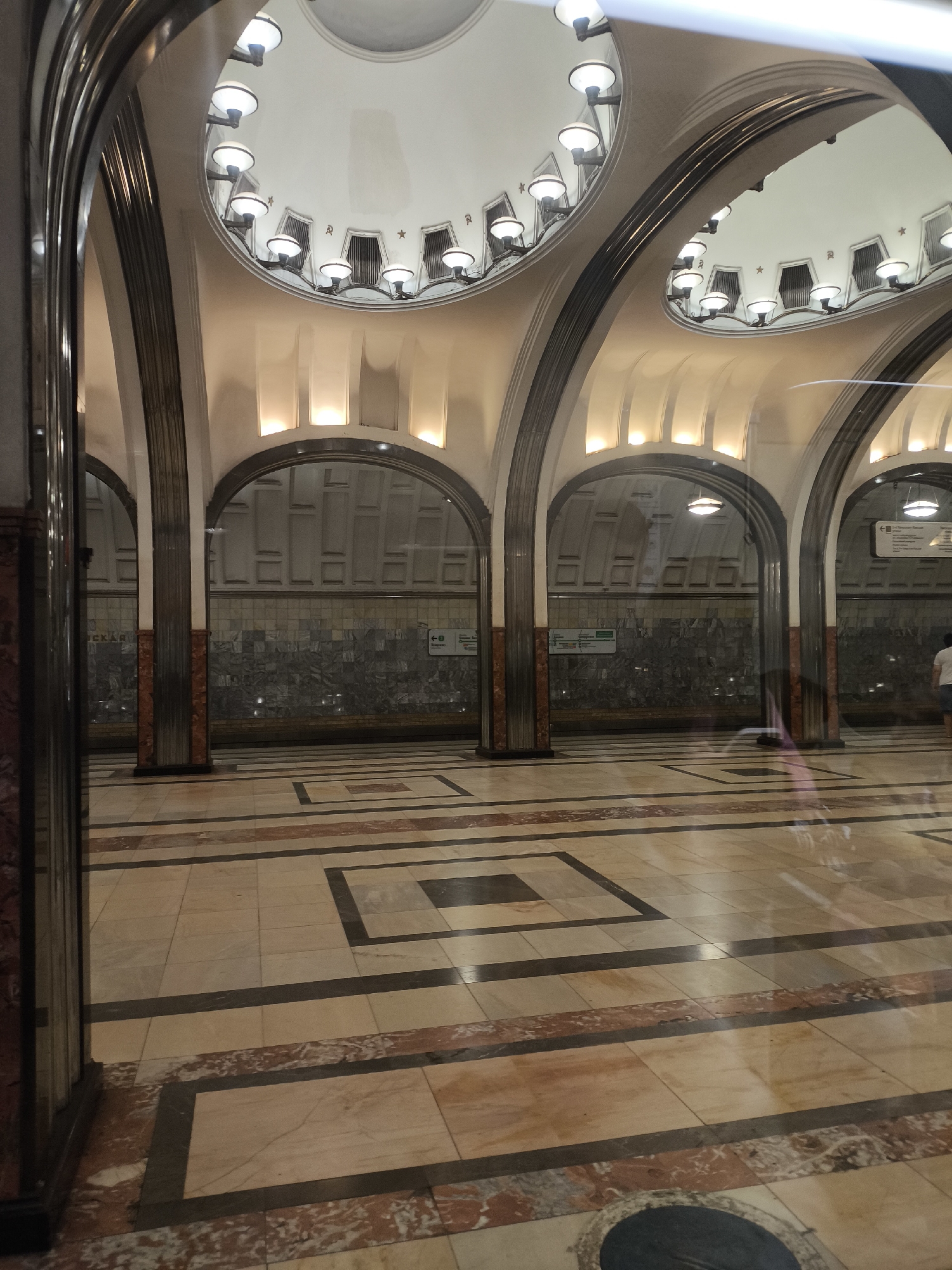 This is a photograph of the interior of a Moscow Metro station, likely Komsomolskaya station, known for its ornate Stalinist architecture. The image captures a section of the station's grand hall, characterized by a series of large arched openings supported by fluted columns. The ceiling features a domed structure with multiple artistic light fixtures, each with a rounded, opaque shade, illuminating the space. The walls visible through the arches are adorned with decorative tiles. The floor is a complex mosaic of polished marble tiles in various colors, including beige, cream, and reddish-brown, arranged in geometric patterns with dark contrasting borders. Reflections are visible on the polished floor and in the glass partitioning the area, indicating a public space with multiple levels or a viewing area. A few people are dimly visible in the background, moving through the station, their forms softened by the reflections and distance. The overall impression is one of opulence and grandeur, typical of the Stalinist era's architectural style in Moscow's underground transport system.