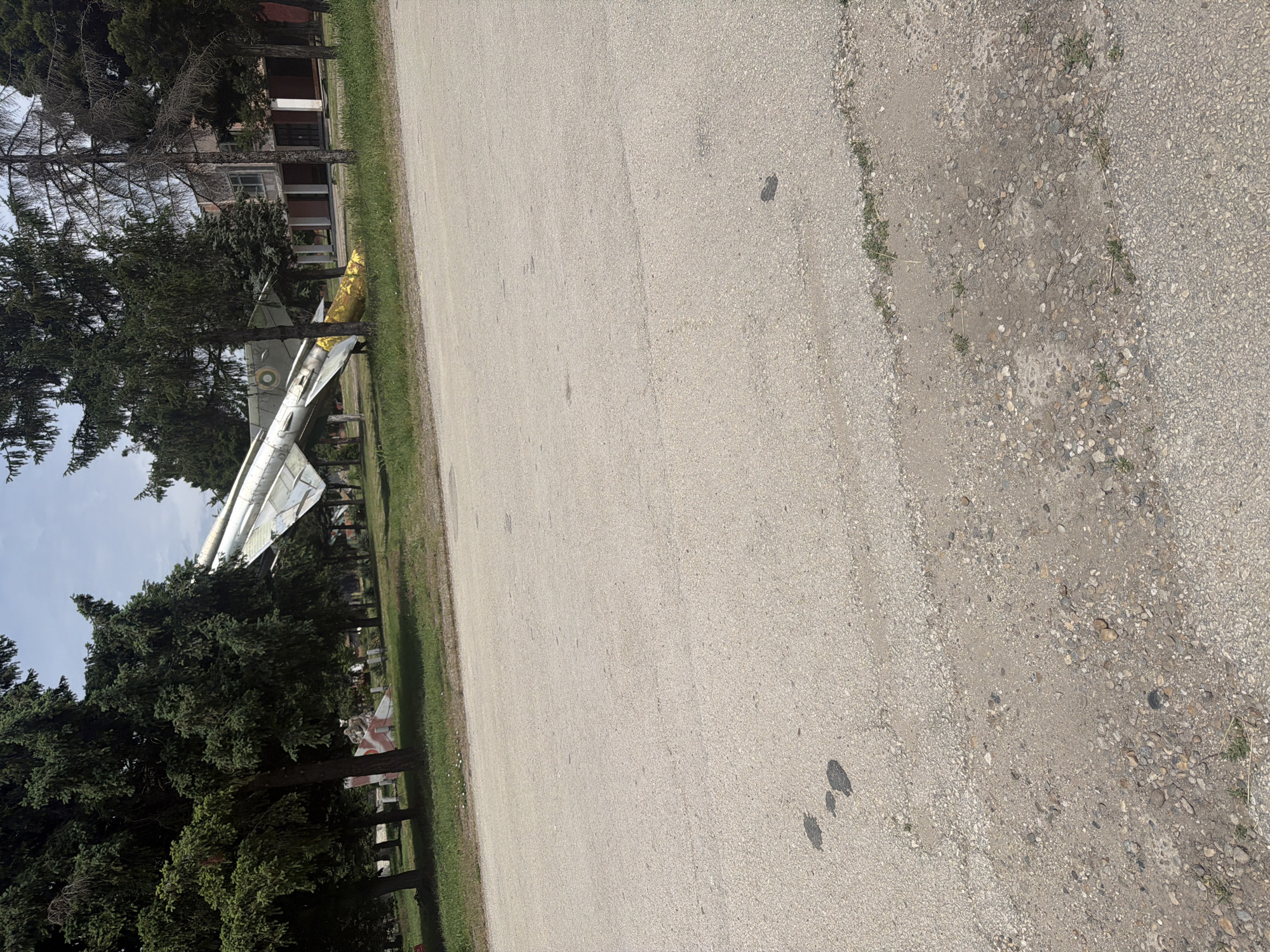 The image shows a paved road or parking area in Dolna Mitropolia, Bulgaria, with a military aircraft, likely a MiG-21, on display. The plane is positioned near trees and grass, with a building visible in the background. The fuselage of the jet is painted in a light color, with the tail section appearing damaged or incomplete and partially wrapped with gold foil. A Red and White flag can be seen under the tree at the end of the frame and appears to be a target. The environment is outdoors, in daylight, with the general setting giving the impression of a static display at a park or monument in the city of Dolna Mitropolia.