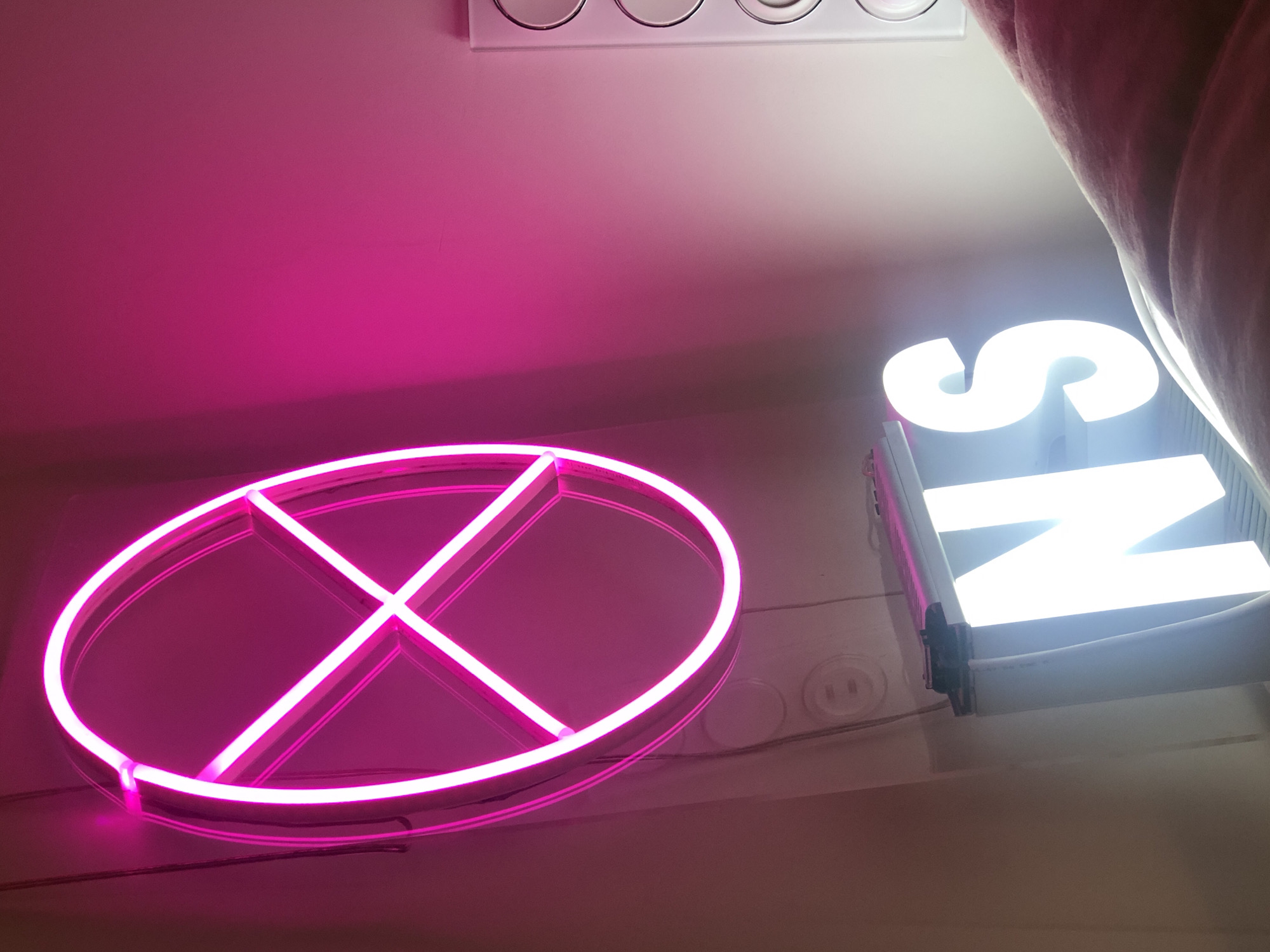 The image displays an indoor scene, primarily illuminated by two distinct artificial light sources. Dominating the upper left portion of the frame is a vibrant, circular neon sign, approximately 60-70 cm in diameter, mounted vertically on a light-colored wall. This sign emits an intense pink-magenta light and features a central cross design within its circular perimeter. Thin dark cables are visible descending from the sign's mounting point.
Below and slightly to the right of the pink neon sign, two bright white illuminated block letters, "N" and "S", are visible, seemingly resting on a flat surface or shelf. These letters emit a cool white light, contrasting with the warm pink glow. The wall behind these light sources is a light, neutral tone, though heavily saturated with pink light on the left and upper sections, transitioning to a lighter, less colored hue towards the right.
On the lower left section of the wall, directly below the pink neon sign, two standard electrical outlets with circular receptacles are visible. On the far right edge of the image, a vertical white panel with a series of four circular depressions, possibly buttons or indicators, is partially visible. The lower right foreground of the image features a textured, upholstered surface exhibiting soft folds and contours, rendered in a muted reddish-brown or dark pink color. No people, specific actions, or identifying city landmarks are present.