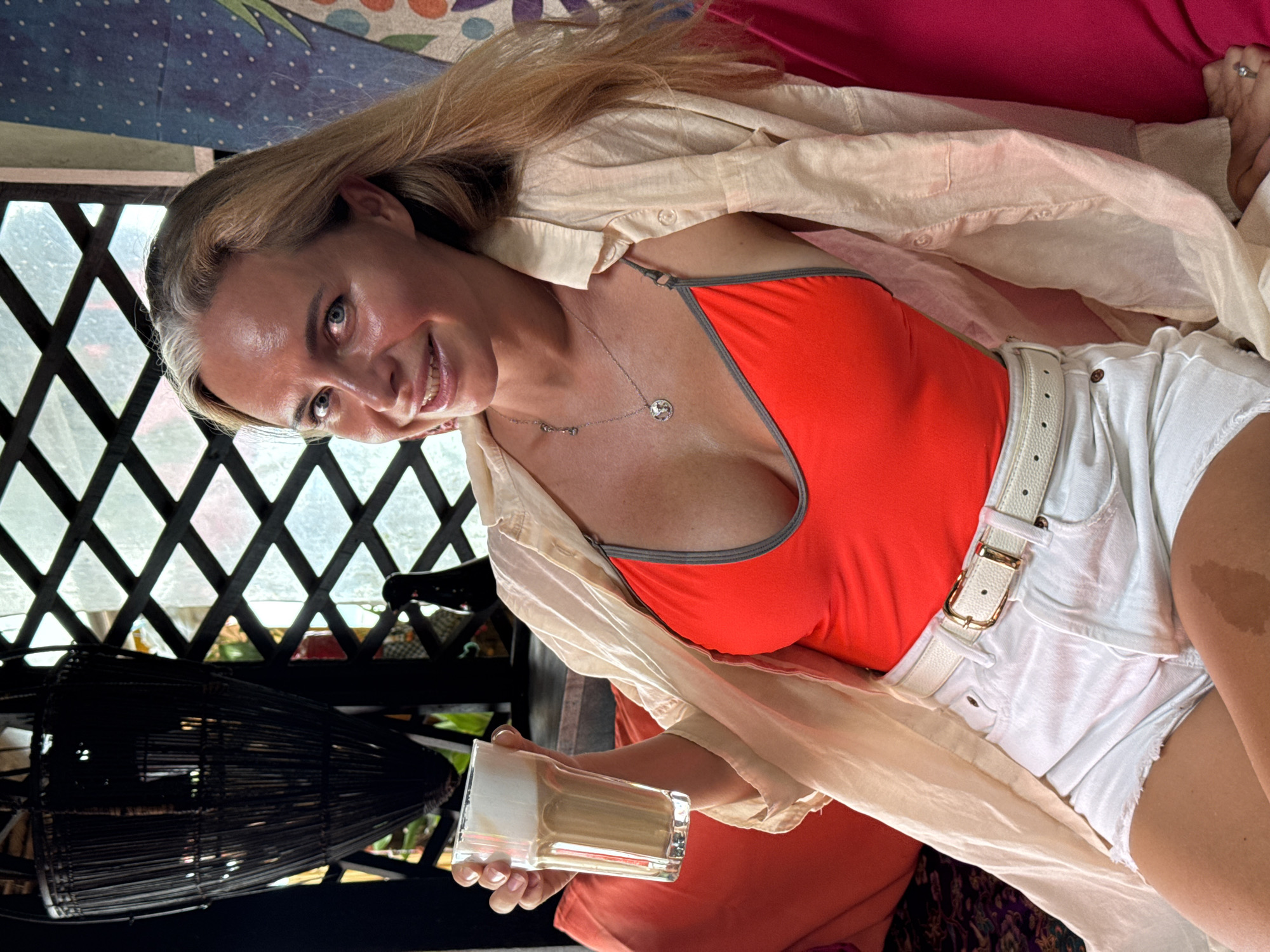 A woman with blonde hair and blue eyes is seated on a couch, holding a glass of iced coffee. She is wearing a red tank top, white denim shorts, and a cream-colored open button-down shirt. She is also wearing a silver necklace and a white belt with a gold buckle. The background appears to be an outdoor seating area with a large lattice window, a dark woven lamp, and colorful cushions. The scene is illuminated by natural light.