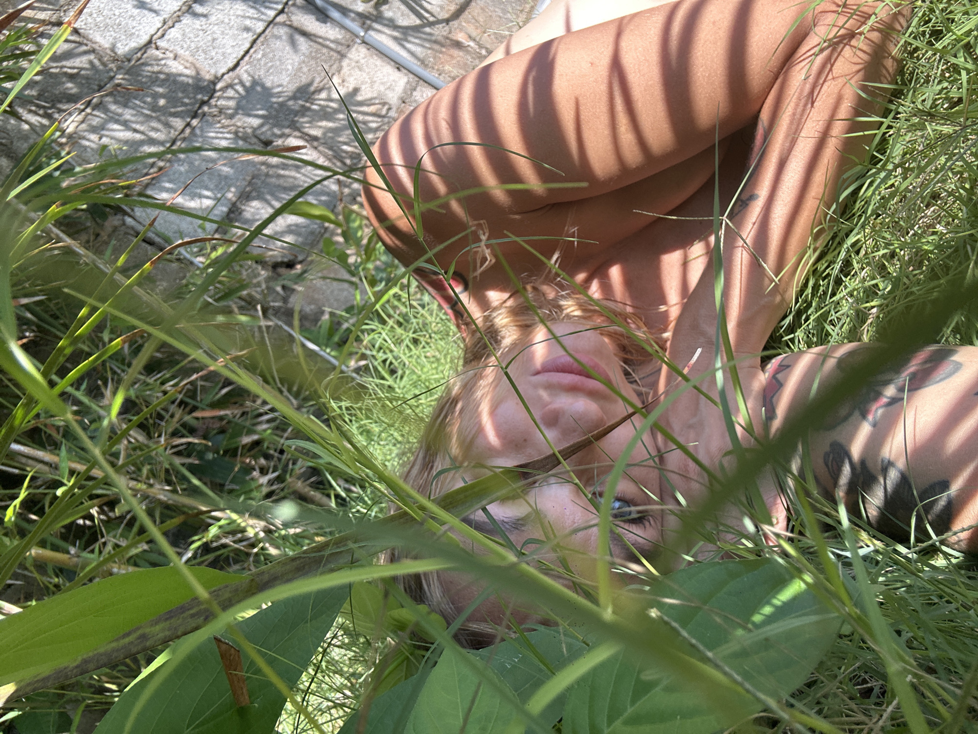 A woman lies on her back in the grass, partially obscured by foliage. Her face, including her eyes and lips, is visible through the blades of grass. Her arms are crossed over her chest, casting shadows. A tattoo is visible on her arm. The ground is a mix of grass, dirt, and paving stones. The location is Mui Ne, Vietnam.