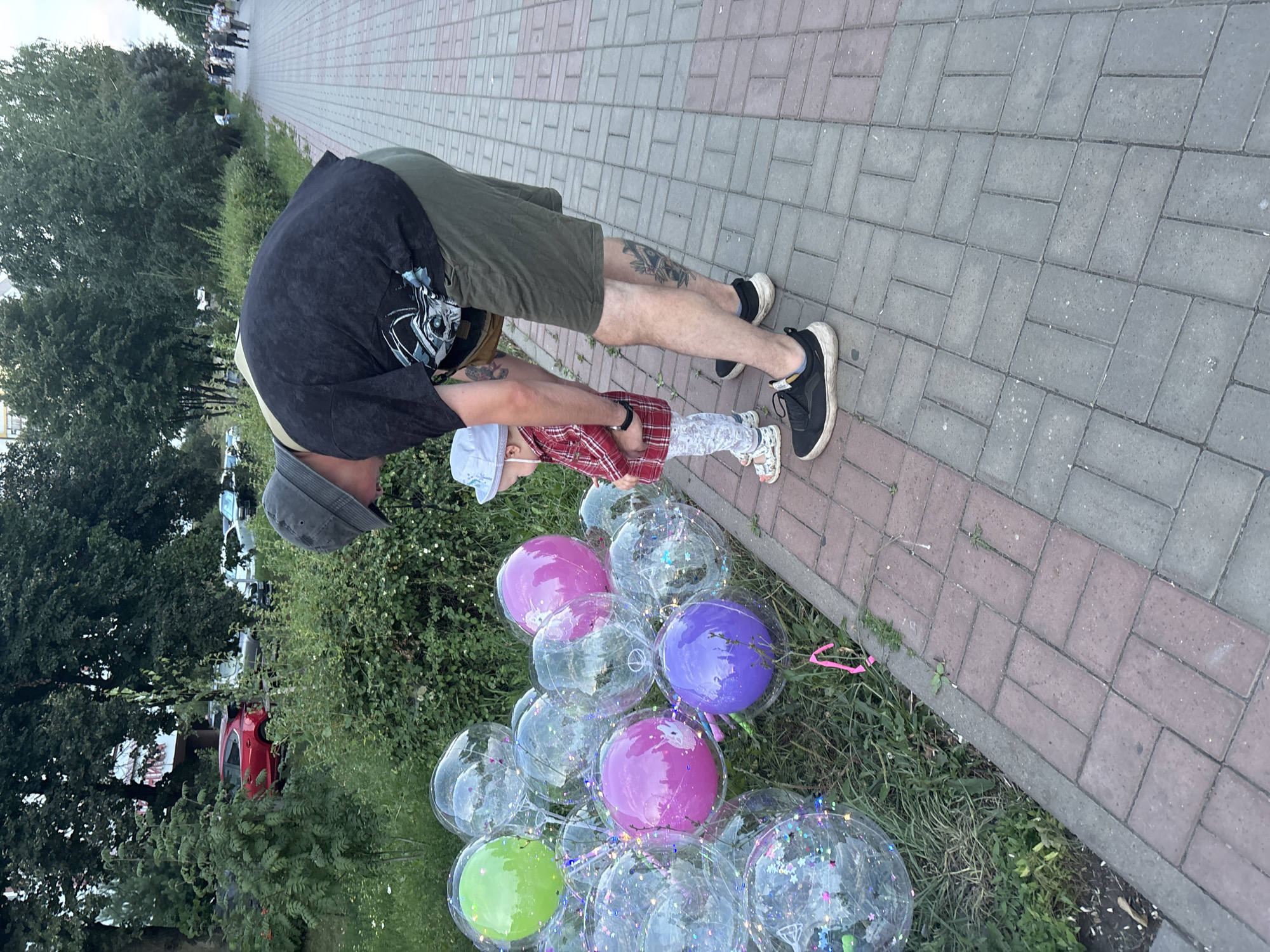 A man wearing a black t-shirt, khaki shorts, and a grey bucket hat is bending down to look at balloons with a young child. The child, wearing a red and white plaid shirt, white pants with a floral pattern, and a light blue hat, is standing next to him. They are on a sidewalk made of red and grey bricks, next to a grassy area with many clear balloons filled with glitter, as well as some colored balloons (green, pink, purple). In the background, trees and cars are visible. This scene is located on a sidewalk in Naberezhnye Chelny, Russia, with a street and more trees in the distance.