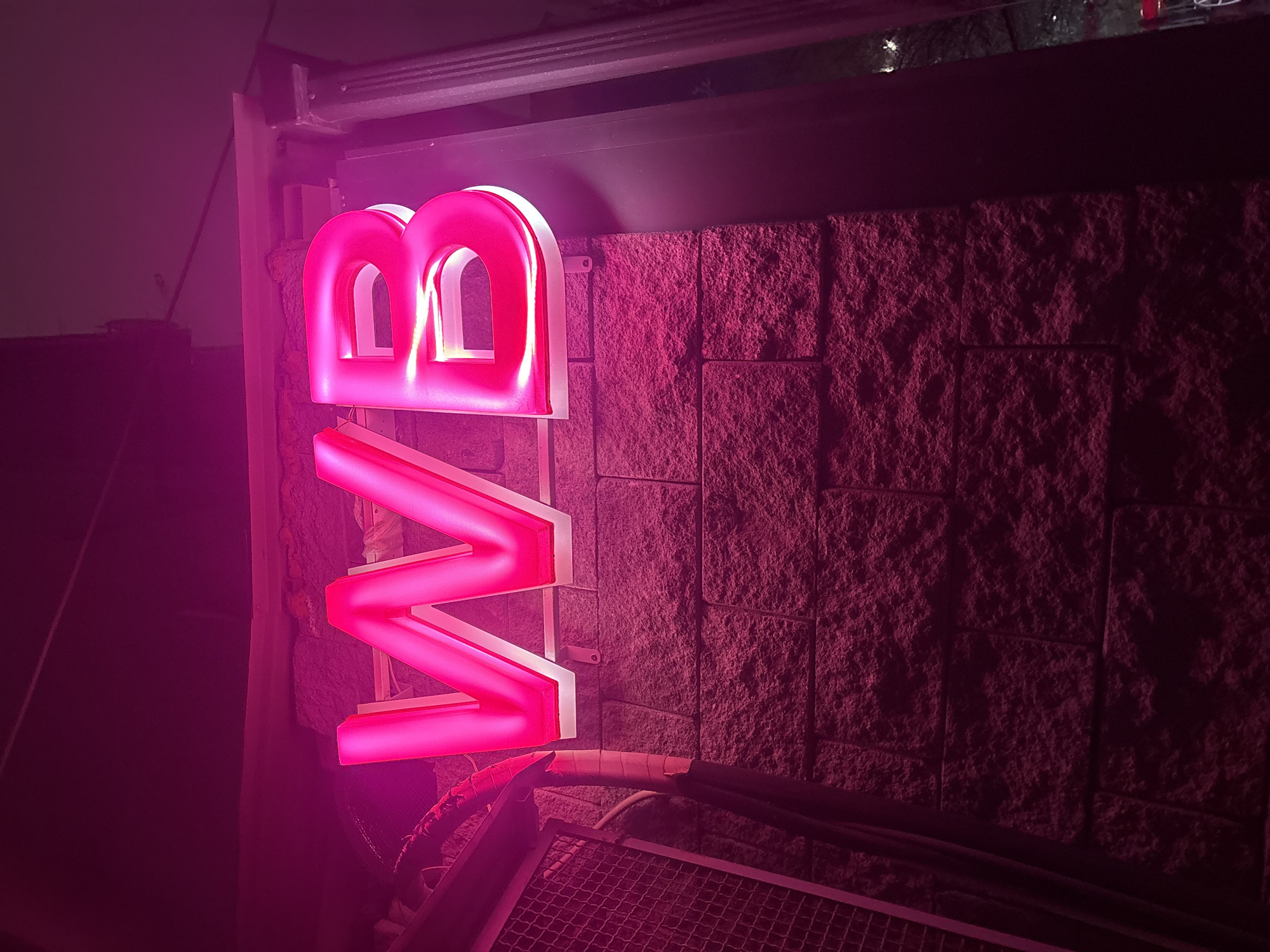 A close-up shot shows a pink neon sign with the letters "VB" mounted on a textured wall. The location is Yerevan, Armenia, based on the letters. The sign is brightly lit, with the pink light reflecting onto the wall behind it. Below the sign, there is a metal grate and some cabling. The overall lighting is low and dramatic, with the sign as the primary light source.