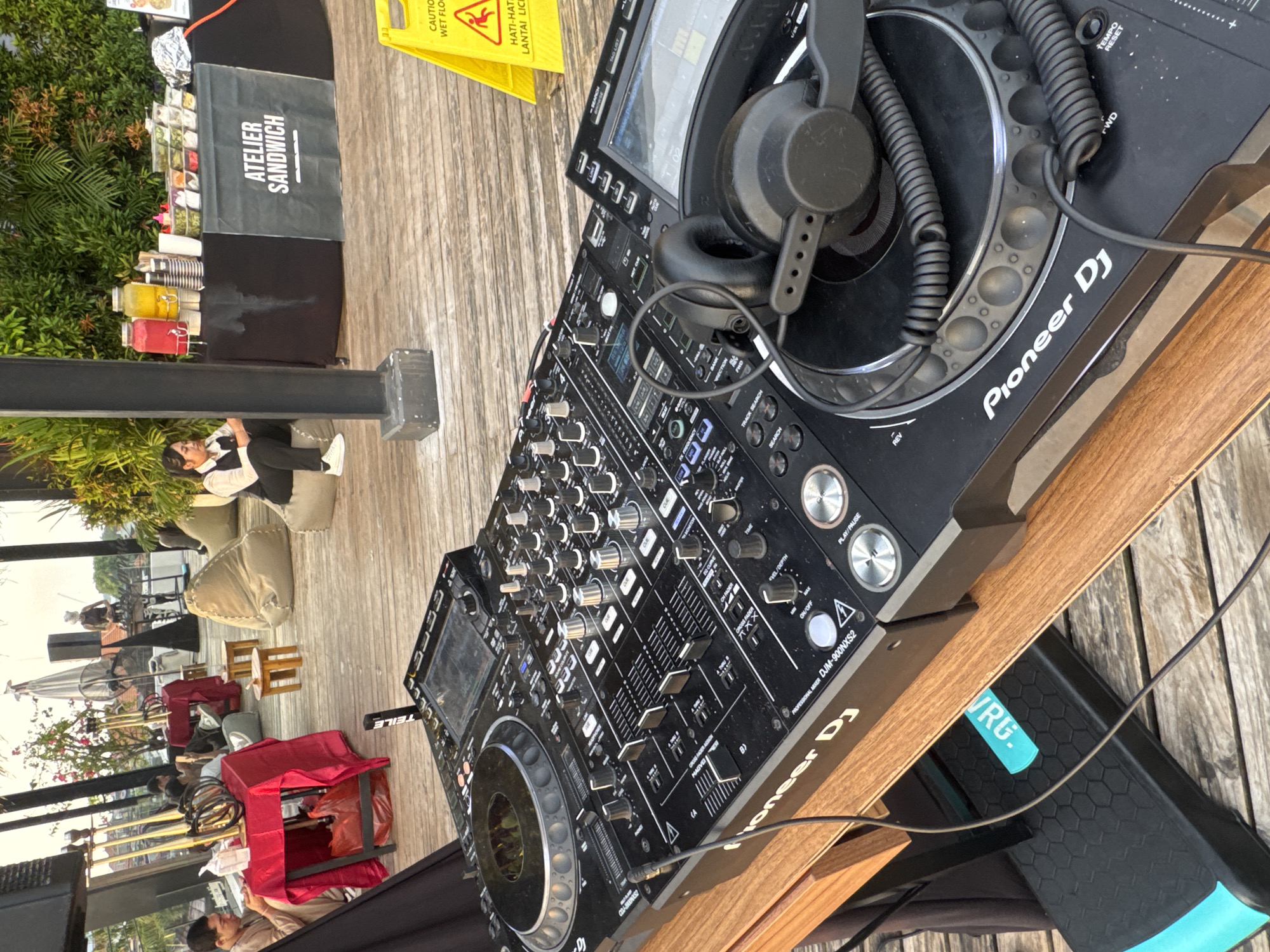 The image shows a close-up of DJ equipment, specifically a Pioneer DJ mixer (DJM-900NXS2) and headphones, set up outdoors on a wooden surface, possibly a table or platform. In the background, there's a glimpse of what appears to be a casual outdoor setting with chairs, tables covered by red fabric, greenery, and patrons. A food stand is visible, advertising "Atelier Sandwich." A yellow caution sign for a wet floor is nearby. Based on the signage "Hati-Hati Lantai Licin" and the label "VRG," combined with the food stand advertising, the location is likely in North Kuta, Indonesia.