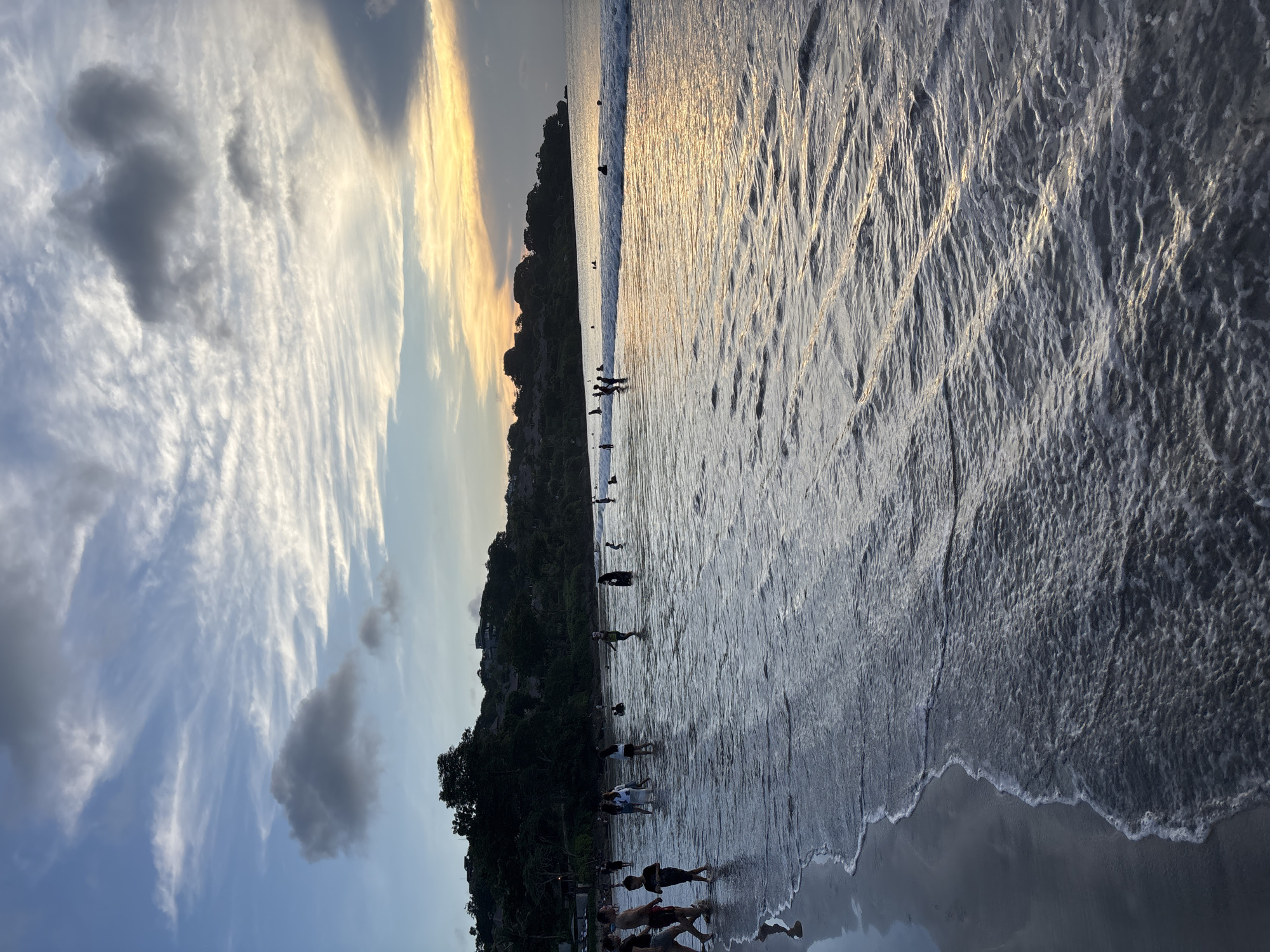 The image depicts a beach scene at sunset. The location is Kuta Selatan, Indonesia. People are present on the beach and in the water, some near the shoreline, others further out. The sun is setting on the horizon, casting a golden reflection on the ocean surface. Clouds are scattered in the sky, displaying a mix of colors from the sunset and blue sky. A line of trees and possibly buildings lines the shoreline. The waves are gently lapping at the shore.