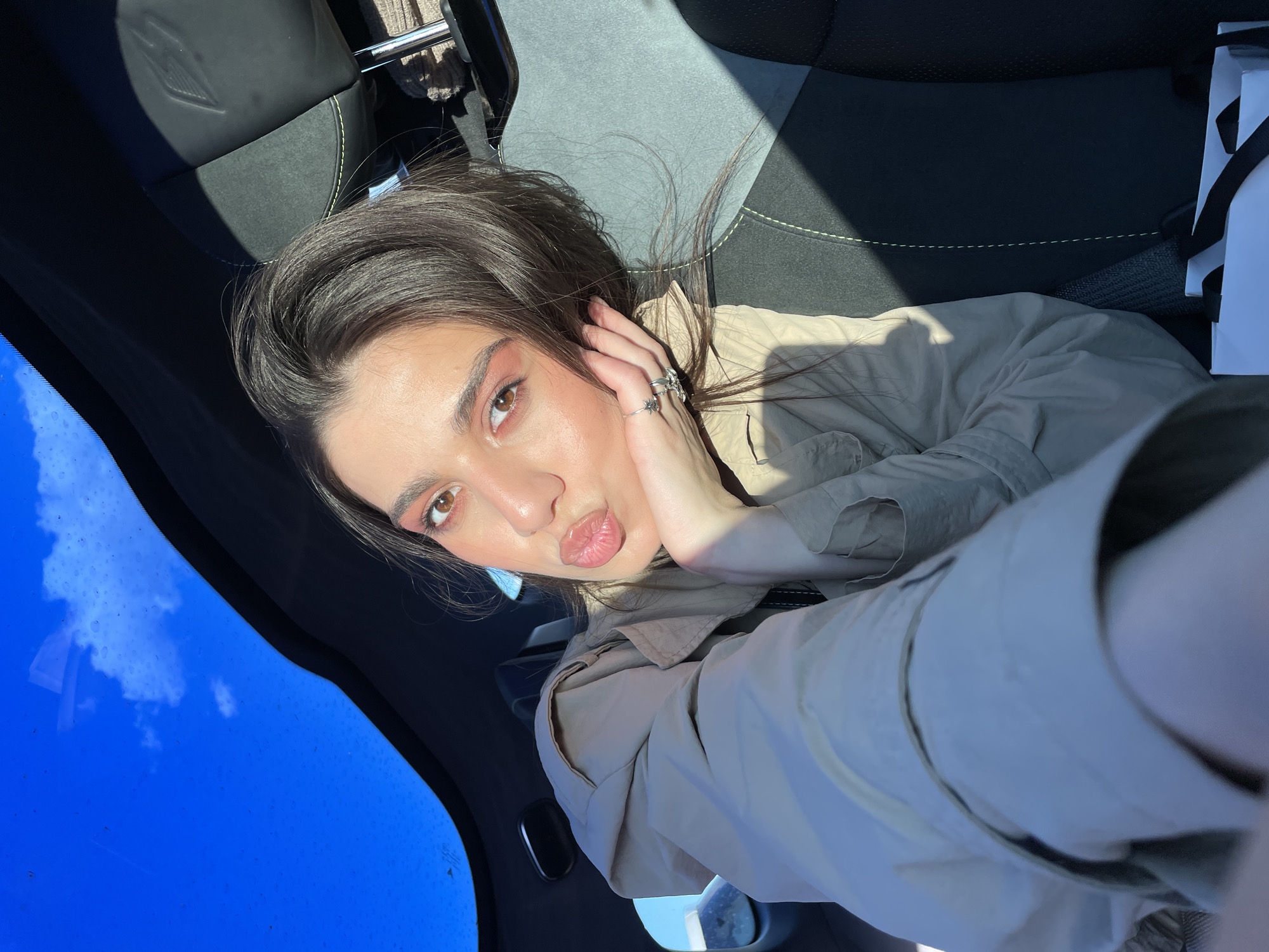 A close-up, high-angle selfie captures a young woman inside a car, looking directly at the camera with a playful "kiss" face. Her lips are puckered, and her left hand is raised to her cheek, displaying several silver rings, including a star-shaped one. She has dark brown hair and is wearing a light-colored (beige or khaki) button-up shirt or jacket. Her makeup features prominent pink/coral eyeshadow and glossy lips.
The setting is the interior of a car, with sunlight streaming in and casting distinct shadows. The car seat behind her is black and grey with light green stitching and a stylized logo, possibly a winged 'A' or 'V', embossed on the headrest. Through the windshield on the left side of the frame, a clear blue sky with some scattered white clouds is visible, indicating daytime and good weather. Small water droplets or dust are present on the windshield. A white shopping bag with black handles is partially visible on the right side of the image, next to the woman. The overall impression is bright and casual. The scene takes place in Apaga, Armenia. No discernible text is visible.
