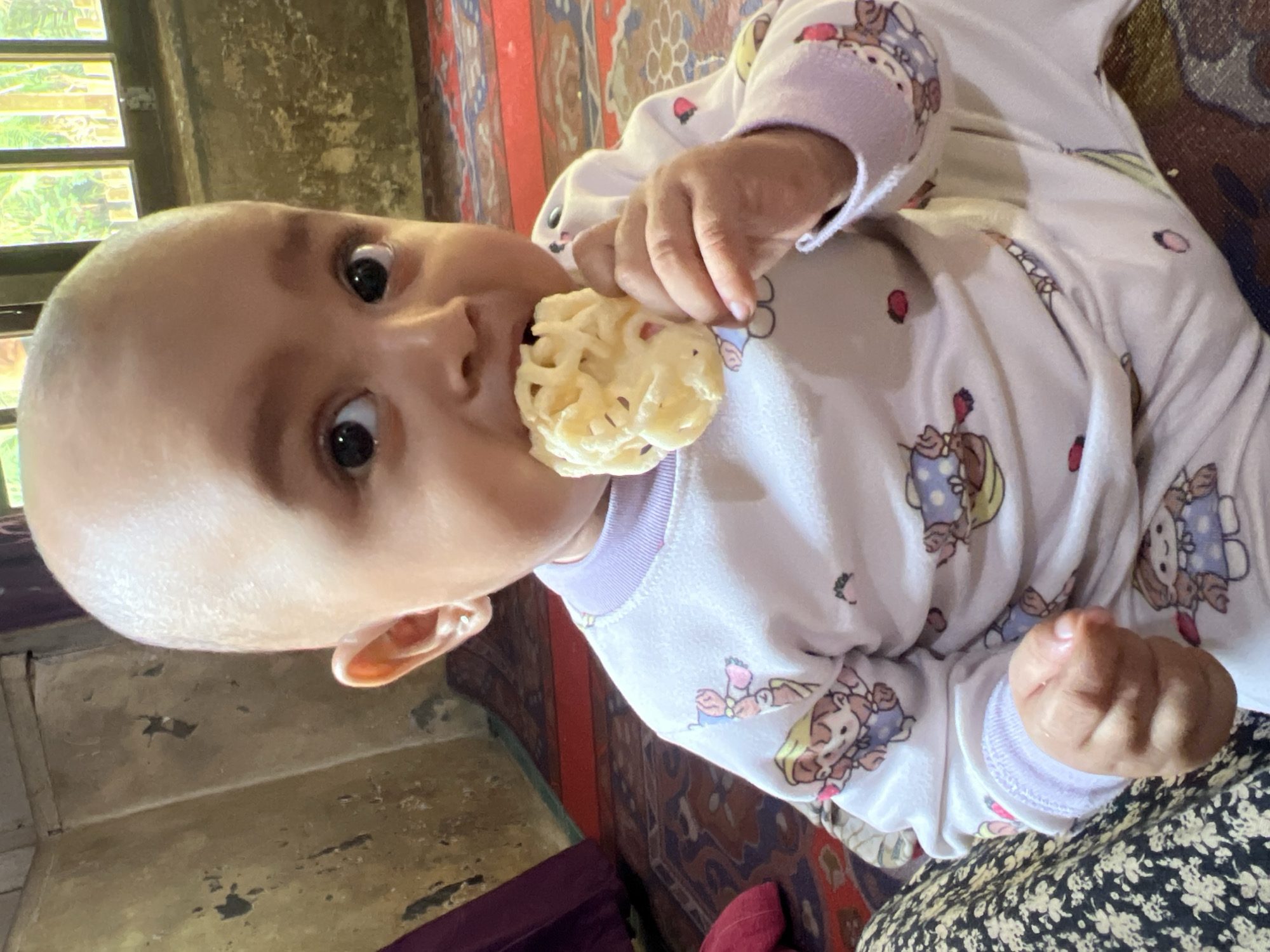 In Kilometer Delapan, Indonesia, a bald baby is seated indoors, wearing a white pajama set adorned with cartoon characters. The baby is holding a white, puffy snack and has it in their mouth, actively eating it. The background shows a textured, aged wall, a window with foliage visible outside, and a patterned rug on the floor. The lighting is natural, casting soft shadows.
