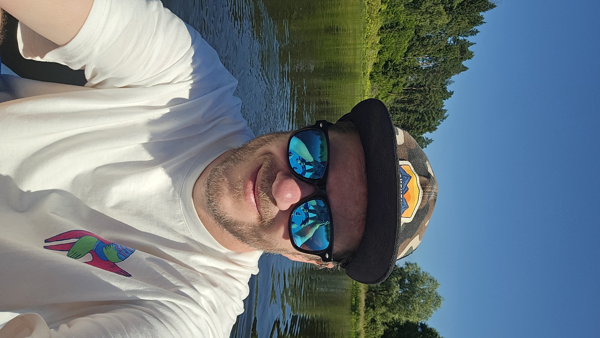 A man with a beard and sunglasses is taking a selfie on a body of water, likely a river or lake, on a sunny day. He is wearing a white t-shirt with a colorful graphic design on the left side. He is also wearing a camouflage baseball cap with an embroidered patch featuring mountains and a sunset. The reflection in his sunglasses shows another person and possibly a kayak or raft. The background consists of trees and a clear blue sky. The location is Voinskaya Chast' Mchs, Russia.