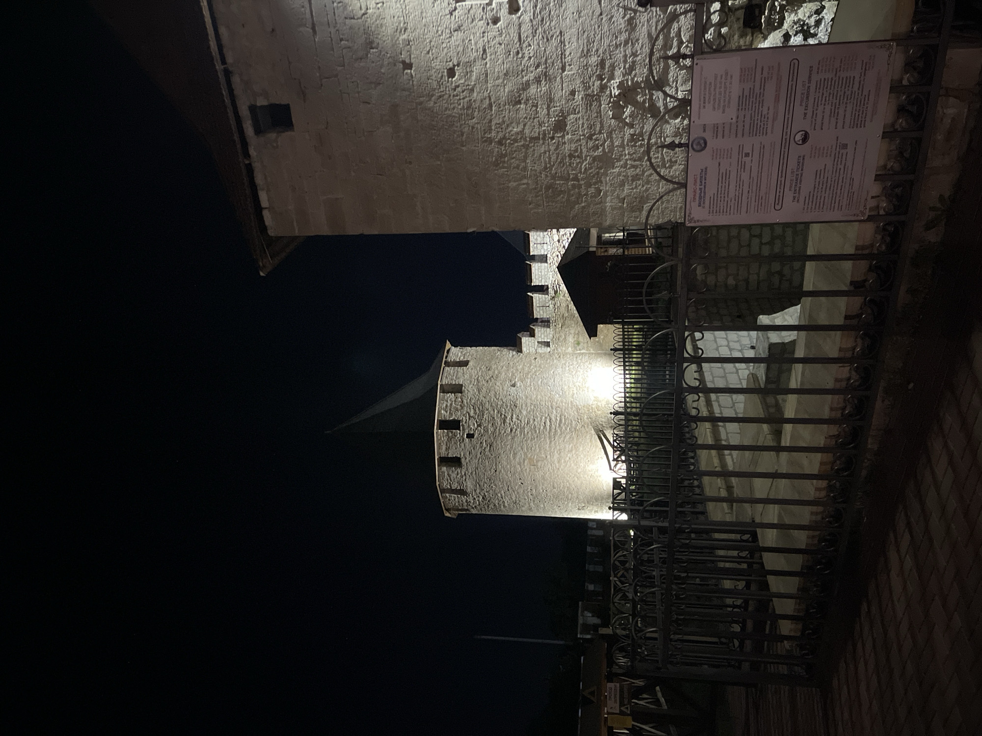 At night, the image shows an illuminated stone tower and a portion of a nearby building behind a dark iron fence. A sign with price lists for entrance tickets and excursion services is affixed to the fence. The location appears to be in Bender, Moldova, as the sign is partly in Russian.