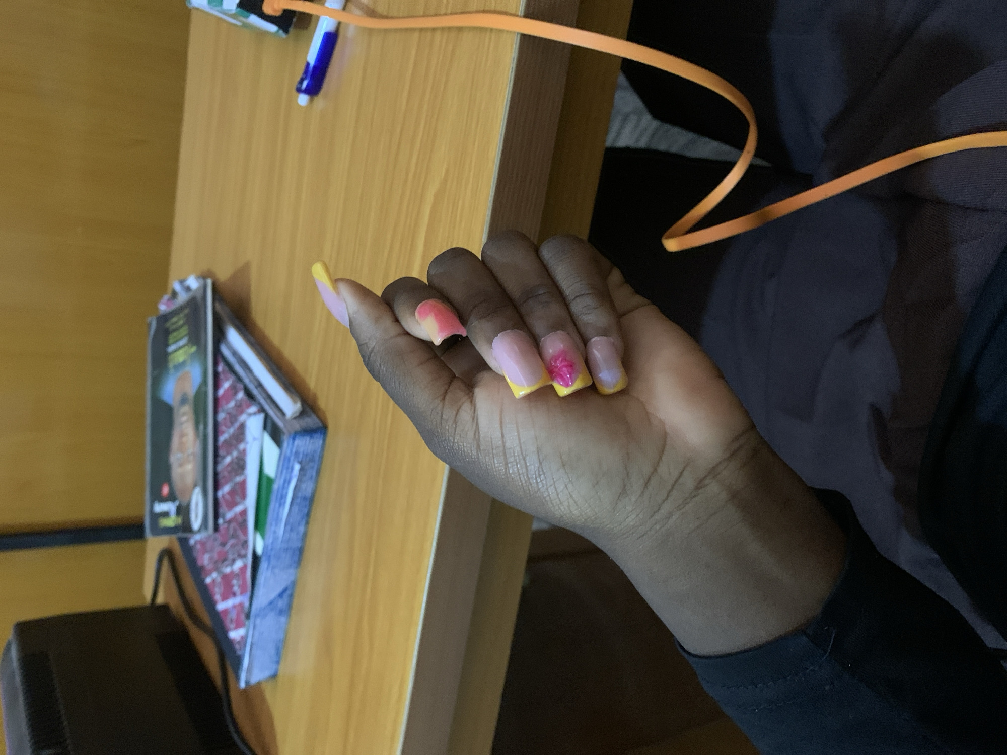 A close-up, medium shot captures a person's left hand with long, acrylic nails, some decorated with pink and yellow ombre and glitter, against a blurred background of a wooden desk. In the background, a black electronic device is visible to the left, and a stack of books or magazines is in the center. To the right, a blue and white pen and an orange cord are also on the desk. The scene is indoors, in an environment that appears to be a study or office.