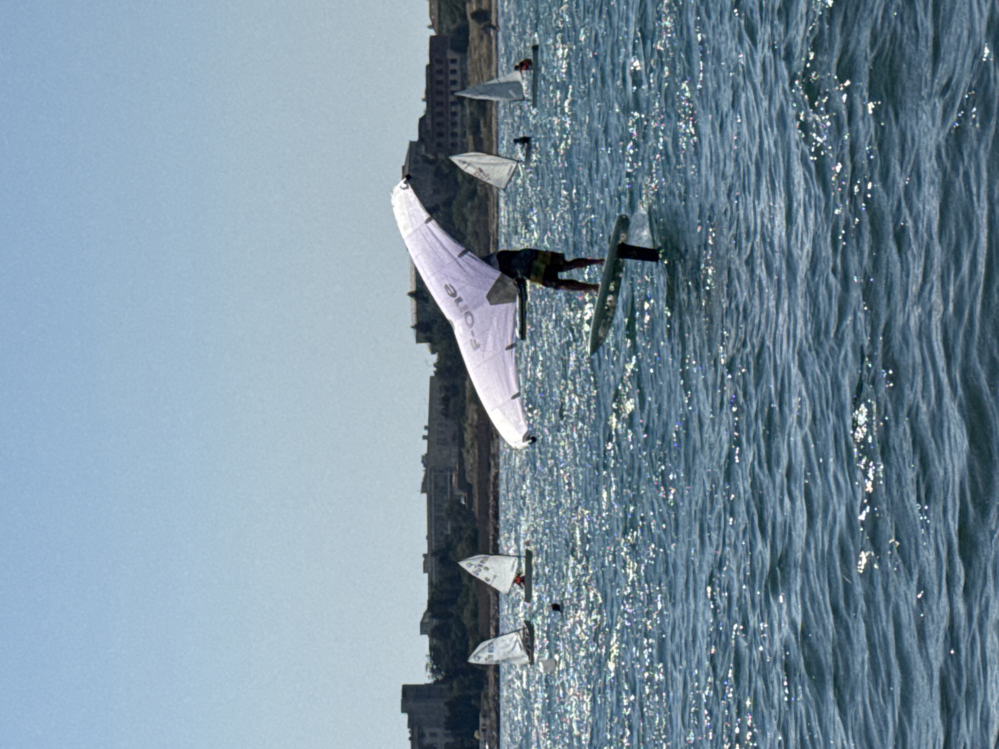An individual is hydrofoiling with a wing in the foreground of a body of water. The wing is predominantly white with a visible "F-ONE" logo. The individual is wearing dark shorts and a yellow garment. Behind the hydrofoiler, several sailboats are visible on the water. The water is choppy and reflects sunlight, creating a sparkling effect. In the background, a coastline features several large, older buildings, consistent with the architecture found in Sevastopol, Ukraine. The sky is clear and light blue.