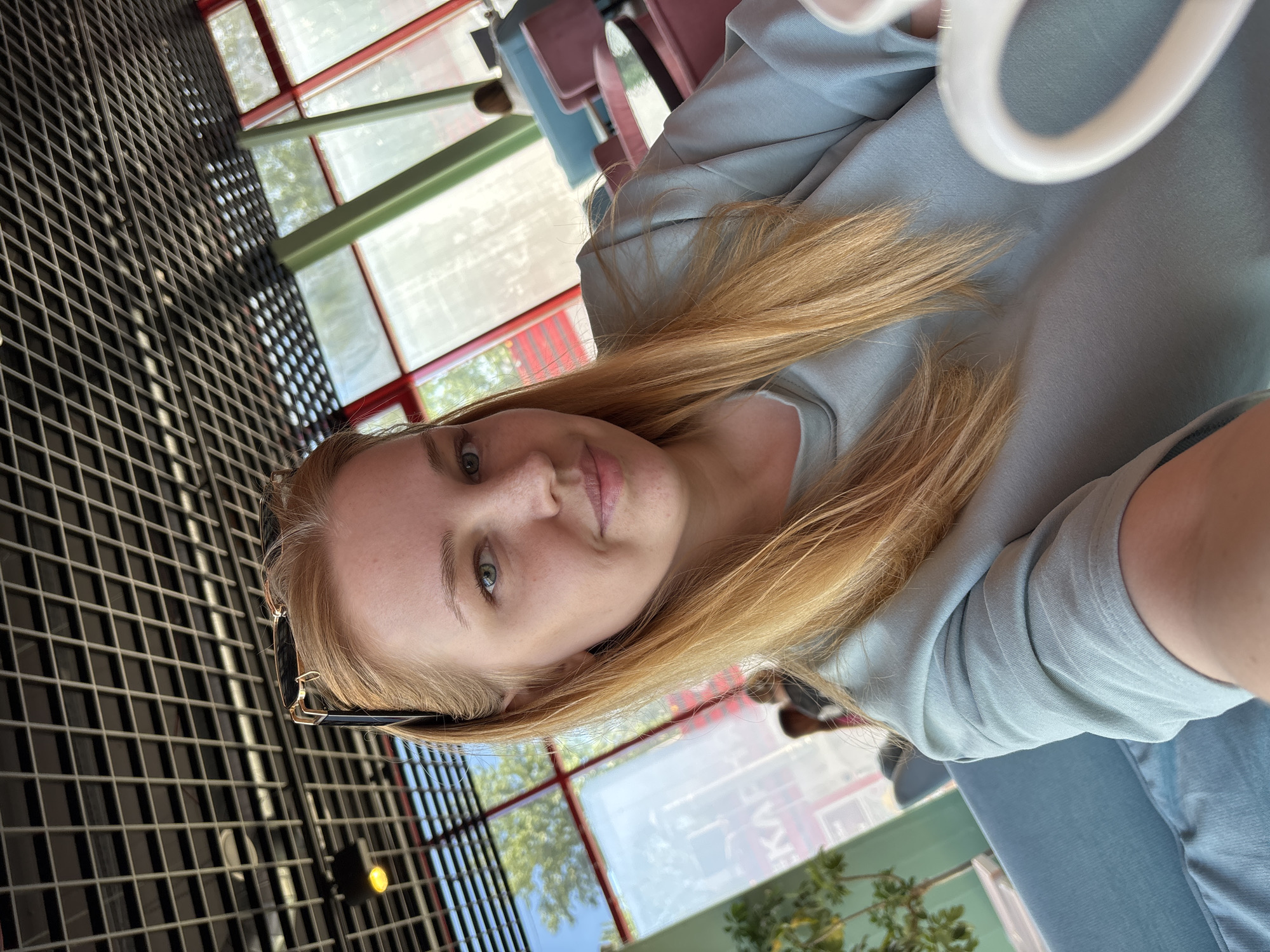 A selfie-style image features a young woman with long, straight, light blonde or light brown hair, wearing a light blue-grey top. Her green-grey eyes look directly at the camera, and she has a neutral to slightly pleasant expression. Sunglasses are perched on top of her head.
The background suggests an indoor setting, possibly a modern cafe or restaurant in Rusanovo, Russia, given the visible Cyrillic text. To the left, a prominent dark metal grid structure, possibly a ceiling or decorative wall element, creates a geometric pattern. Behind the woman, large windows with distinctive red and green frames reveal an outdoor scene with bright daylight, green foliage from trees, and clear sky, indicating it is daytime. Some blurred chairs in shades of pink and blue are partially visible in the midground, further supporting a dining or social venue. On one of the windows, reflections or a sign display Cyrillic text, clearly showing "KAФЕ" (Cafe). The overall image is captured with a diagonal tilt.