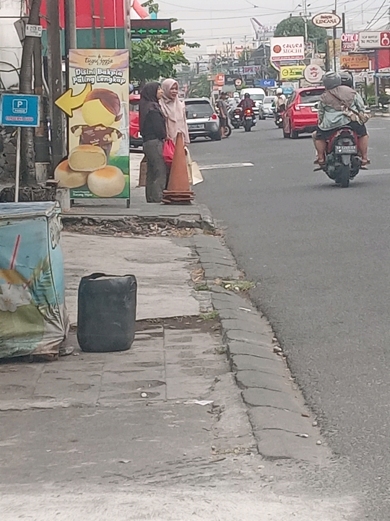 In Depok, Indonesia, on a street with moderate traffic, a scene unfolds near a banner advertising "Tugu Jogja - Bakpia Paling Lengkap". Two women in hijabs stand on the sidewalk. One woman, wearing a black hijab and grey outer garment, holds a red bag. The other woman, in a pale pink hijab and grey outer garment, stands beside her, also holding a red bag. To their right, traffic cones are visible on the sidewalk. A large, dark cylindrical object, possibly a trash bin or water container, sits on the cracked concrete sidewalk near a colorful, tarp-covered object. In the background, the street is busy with vehicles, including cars and motorcycles. A motorcycle with two riders is prominent in the foreground on the right, heading away from the camera. Various storefronts and signs are visible along the street, indicating a commercial area. The overall environment is a typical urban streetscape in Indonesia, with a mix of commercial activity and pedestrian traffic.