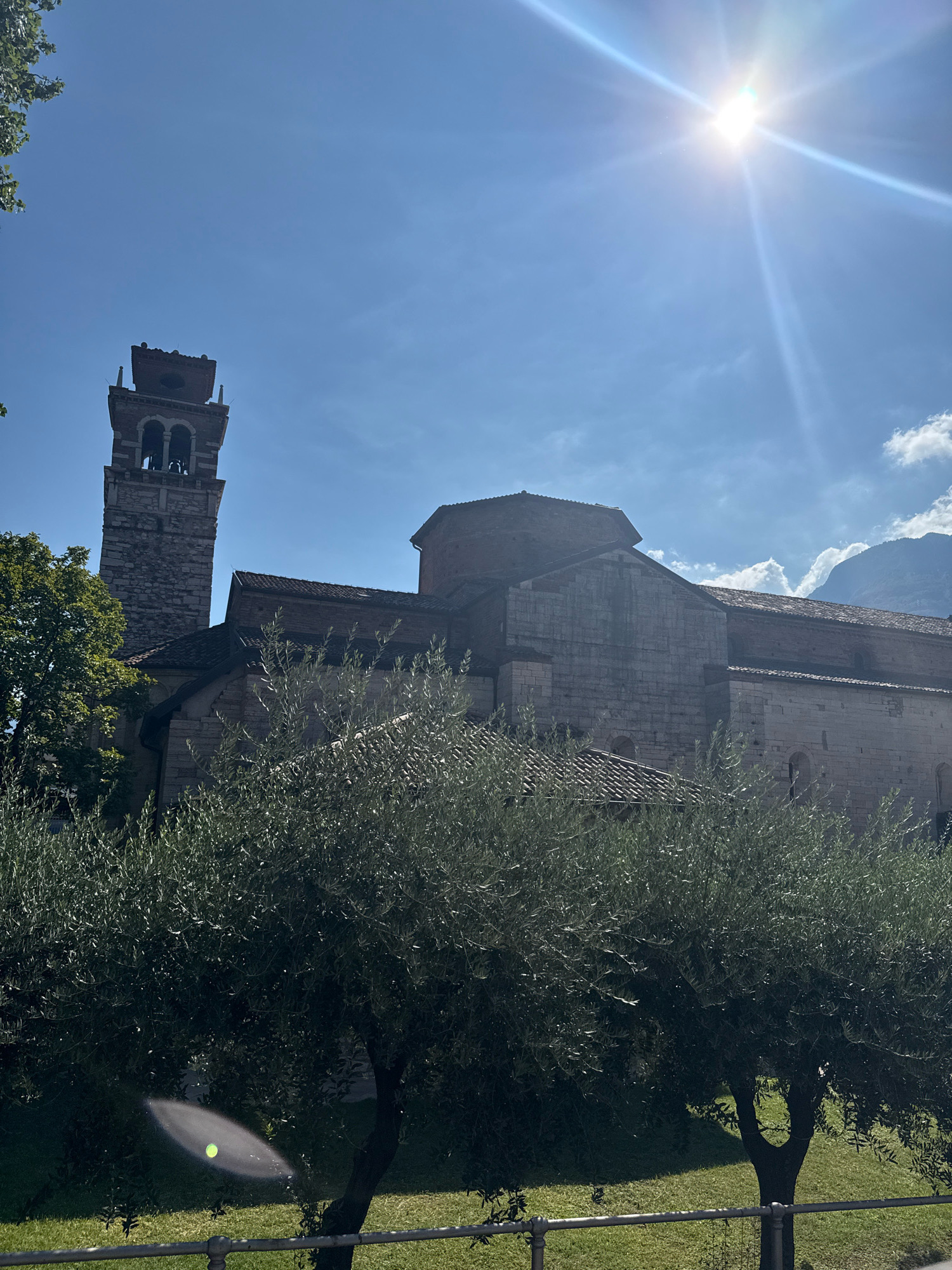 The image depicts the Basilica di San Lorenzo in Trento, Italy, under a clear blue sky with a bright sun. In the foreground, two olive trees with dense foliage partially obscure the view of the ancient stone building. The basilica features a tall, square bell tower constructed of stone with arched openings for bells at the top. The main structure of the basilica is made of large, roughly hewn stone blocks, with a tiled roof. The sun, positioned in the upper right quadrant, creates lens flare and a bright glare across the sky and parts of the image. In the background, the outline of mountains can be seen through the hazy sky. The scene is observed from a ground-level perspective, with a low metal railing visible in the immediate foreground. No people or animals are present in the frame.