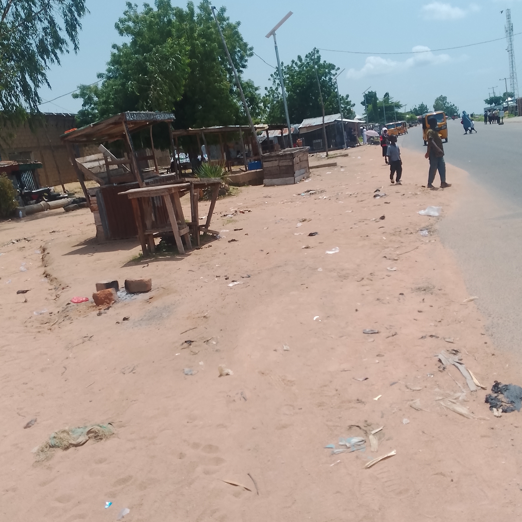 In Jalingo, Nigeria, a dusty, unpaved area adjacent to a paved road features several makeshift wooden stalls, some appearing abandoned. In the foreground, the ground is littered with trash, including plastic bags and debris. Three large stones are arranged in a rudimentary cooking setup. Further down the road, several people are visible, some walking and others gathered. A yellow motorized tricycle, commonly known as a "Keke Napep," is seen in the distance on the road, along with other vehicles. Trees line the background, interspersed with buildings and what appears to be a telecommunications tower. The sky is bright and clear with scattered clouds.