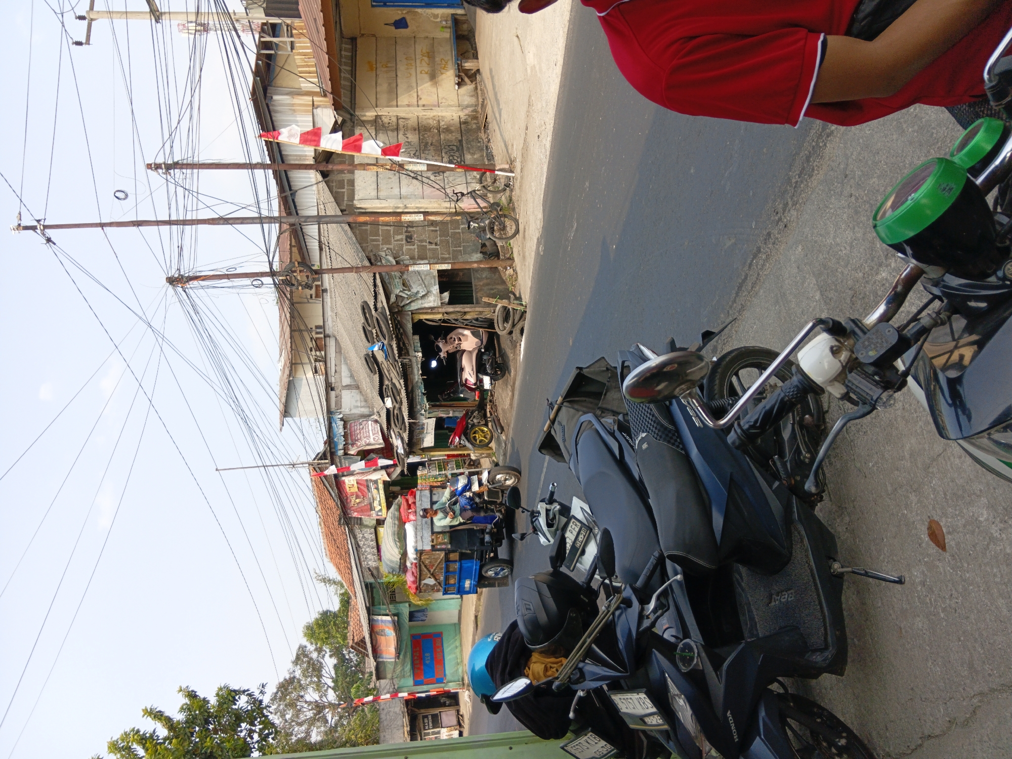 The image depicts a street scene in the city of Bekasi, Indonesia, with a focus on parked motorcycles and a few buildings. In the foreground, several dark-colored scooters are parked on a concrete surface. One scooter has a blue helmet and a black jacket placed on its seat. To the right, a person wearing a red shirt with white trim is partially visible, seated on a motorcycle.
The background reveals a street with a man driving a blue three-wheeled vehicle loaded with goods. Adjacent to the street are several modest buildings with corrugated metal roofs, some appearing to be shops or small businesses. Flags with red and white stripes, resembling the Indonesian flag, are visible on poles outside some of the buildings. Overhead, a dense network of electrical wires crisscrosses the sky. The overall impression is of a bustling, everyday street scene in a densely populated urban area.
