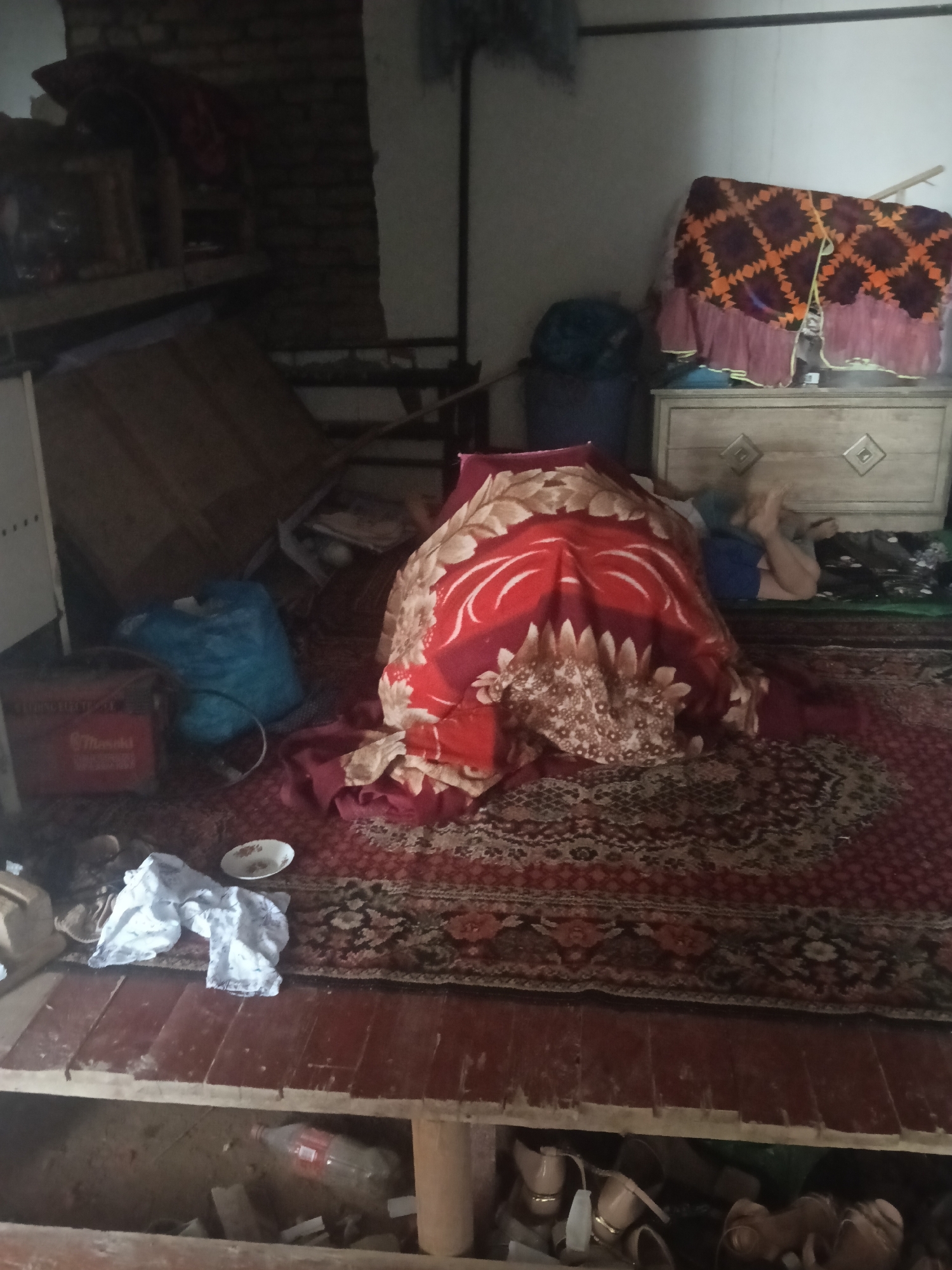 The image depicts an indoor scene, likely a room, with a variety of objects suggesting a lived-in environment. A person, whose legs and lower torso are visible, is lying down on a red and gold patterned rug, covered by a red blanket with a floral design. The person is wearing blue shorts. To the right of the person is a light-colored wooden dresser with diamond-shaped decorative inlays. Above the dresser, a patterned fabric with a diamond motif in orange, brown, and black, possibly a blanket or tapestry, is draped over a support.
In the foreground, the floor is made of wooden planks, some of which appear to be missing or damaged, revealing a space below containing a plastic bottle and other debris. A red box with text is visible to the left, next to a large blue bag. A small white bowl with some residue sits on the rug. White cloth, possibly laundry, is also present on the rug. The walls in the background are made of brick and plaster. The overall impression is one of a somewhat cluttered and modest living space. The mention of Altinkul, Uzbekistan, places this scene within that geographical context.