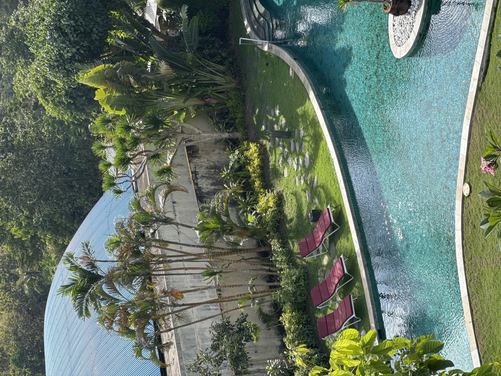 The image depicts an outdoor scene in Kuta Selatan, Indonesia, featuring a long, narrow swimming pool with turquoise water. The pool is lined with a grassy area and several red lounge chairs are placed on the grass beside it. Adjacent to the grass, there is dense tropical foliage and a building with a corrugated metal roof. The scene is captured from an elevated perspective, likely from a higher floor of a nearby building.