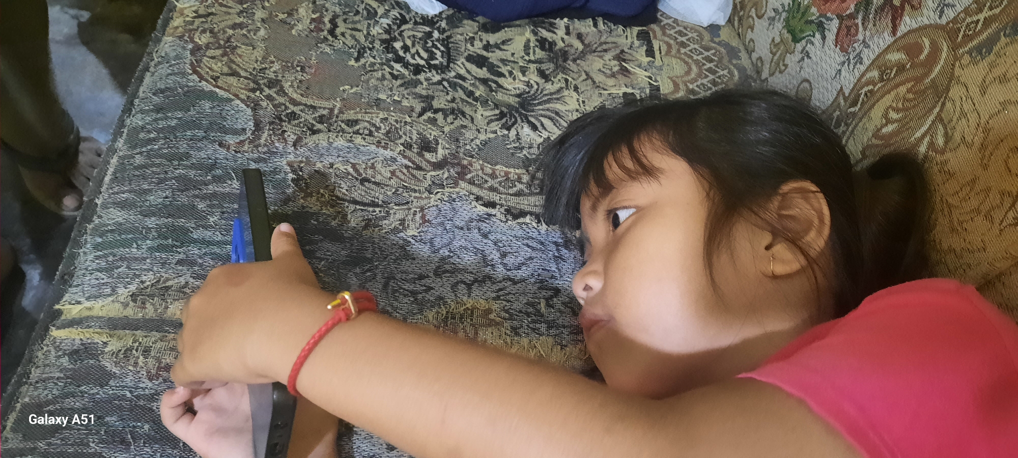 A young girl, likely of Southeast Asian descent, is lying on a patterned sofa, engrossed in a smartphone. She is viewed from the side, with her face turned towards the device, her eyes wide with attention. A red braided bracelet adorns her left wrist. Her hair is dark and straight, pulled back from her face. She is wearing a vibrant pink top.
The sofa has a textured, ornate fabric with floral motifs in earthy tones. The background of the image is dimly lit, suggesting an indoor setting, possibly a home, with a hint of a concrete floor visible on the left. The overall mood is one of quiet focus, with the girl fully absorbed in her digital activity. A portion of a dark object, potentially the smartphone or a case, is held in her hand. The lighting is soft, casting a shadow across her face, highlighting her profile. The scene appears to be captured in natural light, possibly during the day, though the interior setting makes it difficult to ascertain the exact time. The "Galaxy A51" watermark is visible in the bottom left corner, indicating the device used to capture the image.