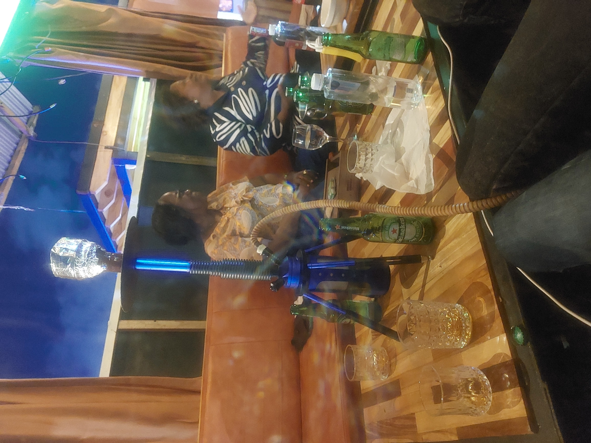 The image shows an indoor scene, likely a lounge or bar, in Dar es-Salaam, Tanzania. Two women are seated. One is wearing a patterned black and white shirt, and the other is wearing a floral top. They are positioned near a table that is crowded with objects including several Heineken beer bottles, various clear glasses (some appear empty, others may contain liquid), and a white napkin. A hookah, distinctively blue with a silver foil-wrapped bowl, is located closer to the foreground of the image. The seating appears to be cushioned. The lighting suggests evening or nighttime. The environment has a casual, social atmosphere. The background has elements of a building's structure visible.
