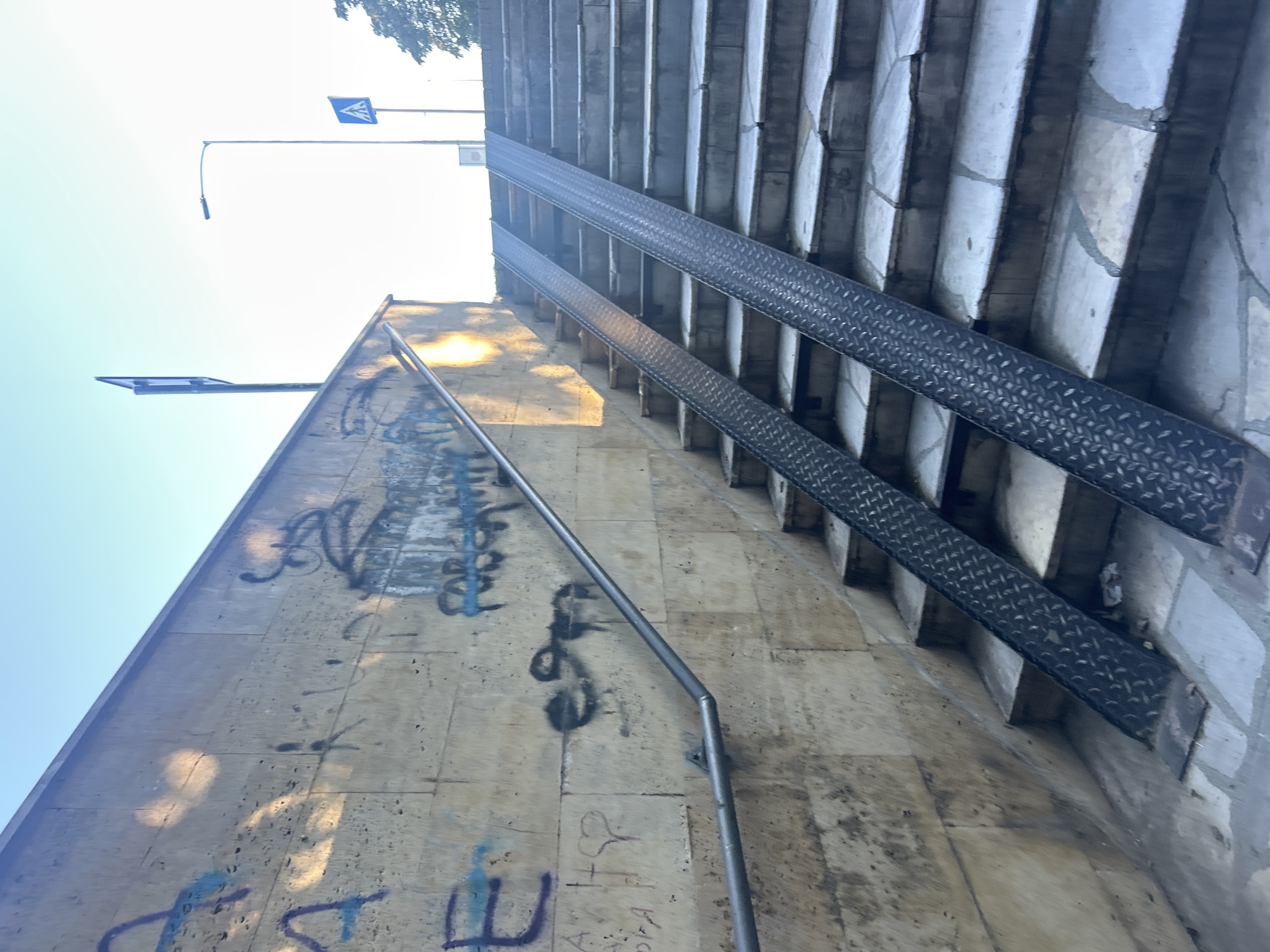 This is a street-level, outdoor, eye-level shot of a set of stairs with two metal ramps installed. The stairs are made of light-colored stone blocks, some of which appear cracked or worn. The ramps are made of dark, textured metal. The wall to the left of the stairs is made of the same light-colored stone and is covered in graffiti in various colors including blue, black, and purple. A metal handrail runs along the left side of the stairs and wall. The scene is bathed in the warm light of the sun, casting shadows. In the background, there is a street lamp and two pedestrian crossing signs. The sky is clear and blue. The overall impression is one of urban infrastructure, possibly designed to improve accessibility.
