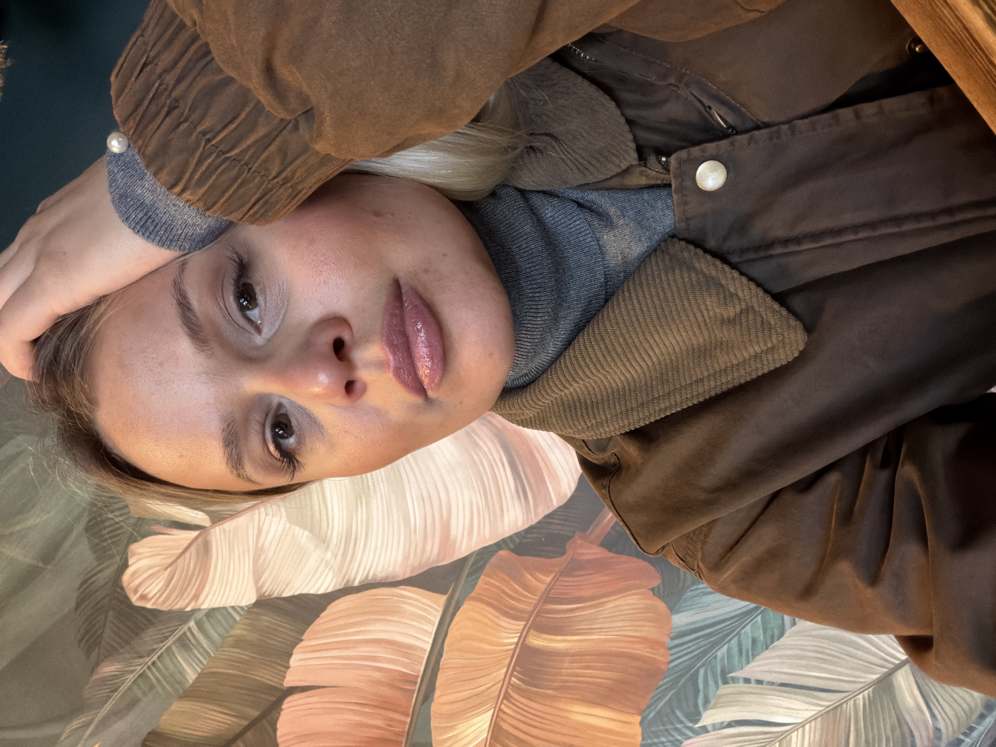 A blonde woman with light skin and glossy lips is positioned in the center of the frame. She is wearing a gray turtleneck sweater and a brown jacket. Her right hand rests on her head, adorned with a pearl bracelet. The background features a patterned surface with large, stylized leaves in shades of peach, green, and white. The lighting is bright and appears to be artificial. The scene is indoors and appears to be a casual setting. There is no information to verify this image was taken in Sarapul, Russia.