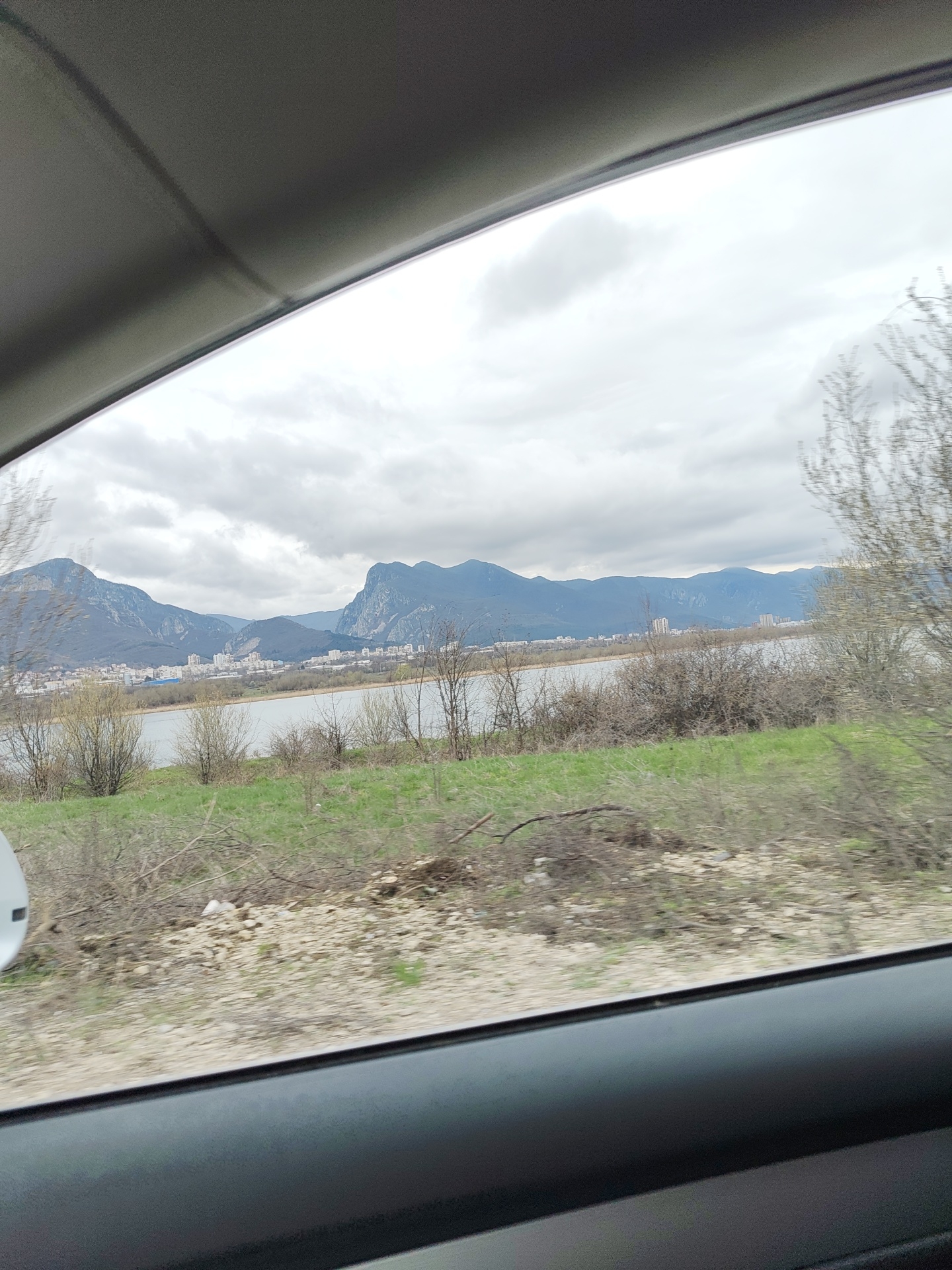 The image depicts a landscape viewed through the window of a moving vehicle. In the background, a body of water stretches across the scene, with a city nestled along its shore. Behind the city rise a series of mountains. The foreground shows a stretch of land covered with low vegetation, including grass and bushes. The overall scene suggests a location in or near Vratsa, Bulgaria, based on the characteristic mountain range. The sky is overcast. The vehicle's interior framing the view indicates the shot was taken from inside the car. The motion blur suggests the vehicle is moving. There are no visible people in the scene.
