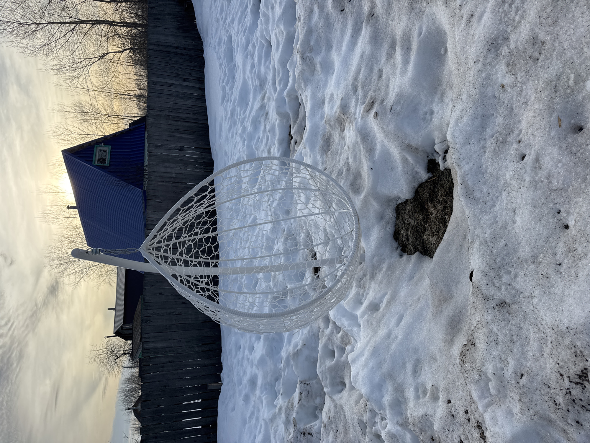 An outdoor scene in Chaikovsky, Russia, depicts a white, egg-shaped hanging chair suspended from a matching stand, positioned slightly left of center in the foreground. The ground is covered by uneven, melting snow, exhibiting visible dirt and brown earth patches, particularly a prominent circular area directly beneath and slightly to the right of the chair. In the mid-ground, a weathered, dark wooden picket fence spans horizontally across the frame. Beyond the fence, several buildings are visible, including a structure with a steep, bright blue corrugated metal roof and a small, green-framed attic window on the right. Bare deciduous trees are interspersed throughout the background, indicative of a late winter or early spring setting. The sky is partially cloudy, with the sun visible through the clouds near the horizon on the right, casting a soft, warm light across the scene. No individuals are present.