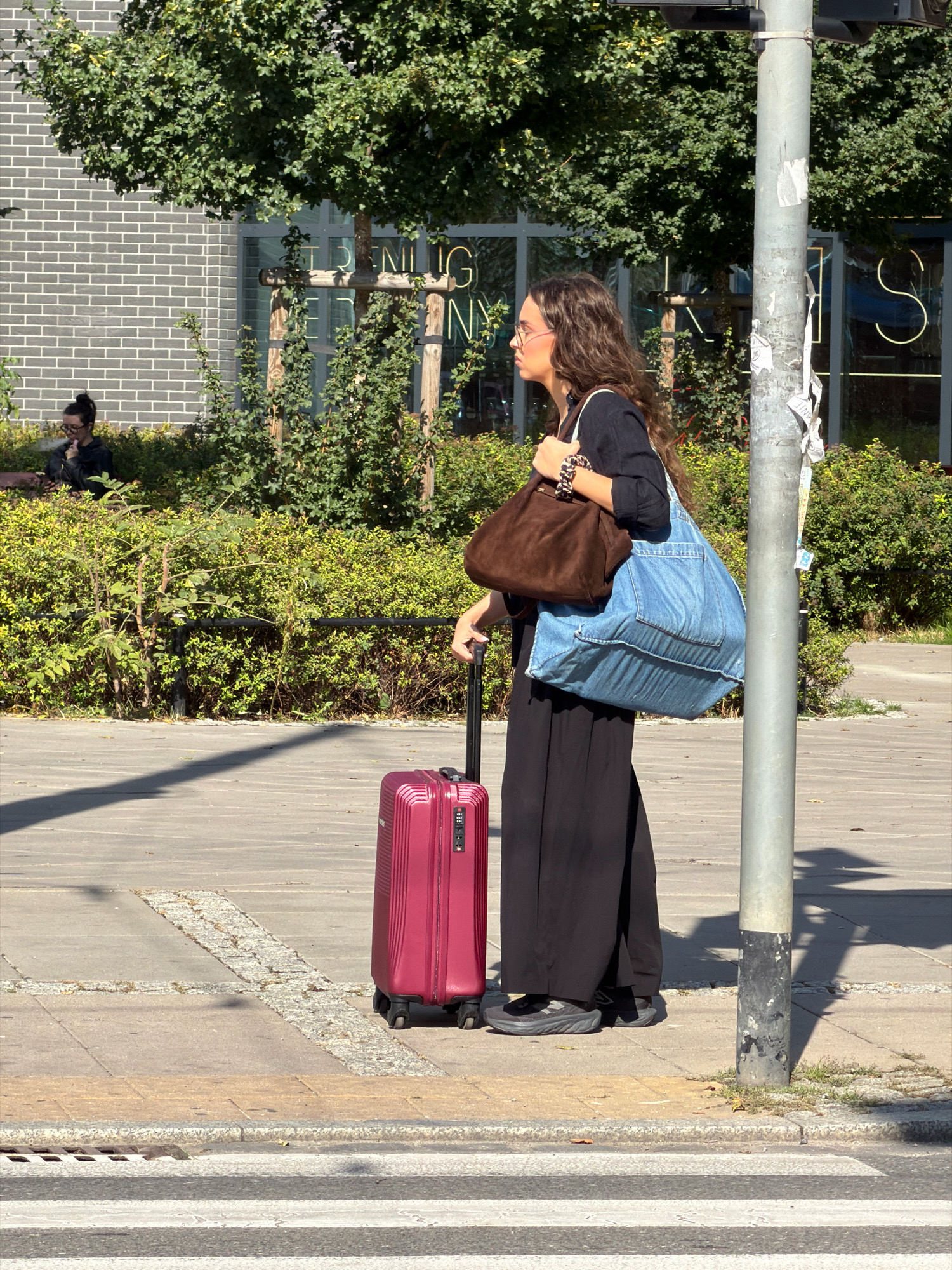 A woman stands on a sidewalk in Wrocław, Poland, holding a suitcase and two large bags. She is wearing a long black dress, dark sneakers, and glasses, with long brown curly hair. A pink suitcase with a combination lock is beside her. She carries a brown suede bag over her left shoulder and a large blue denim tote bag in her right arm. In the background, a brick building with large windows and green trees can be seen. A streetlamp is to her right. The sidewalk is paved with grey stones and yellow concrete curbs, with a crosswalk visible in the foreground. Another person is seated on a bench in the distance, smoking.