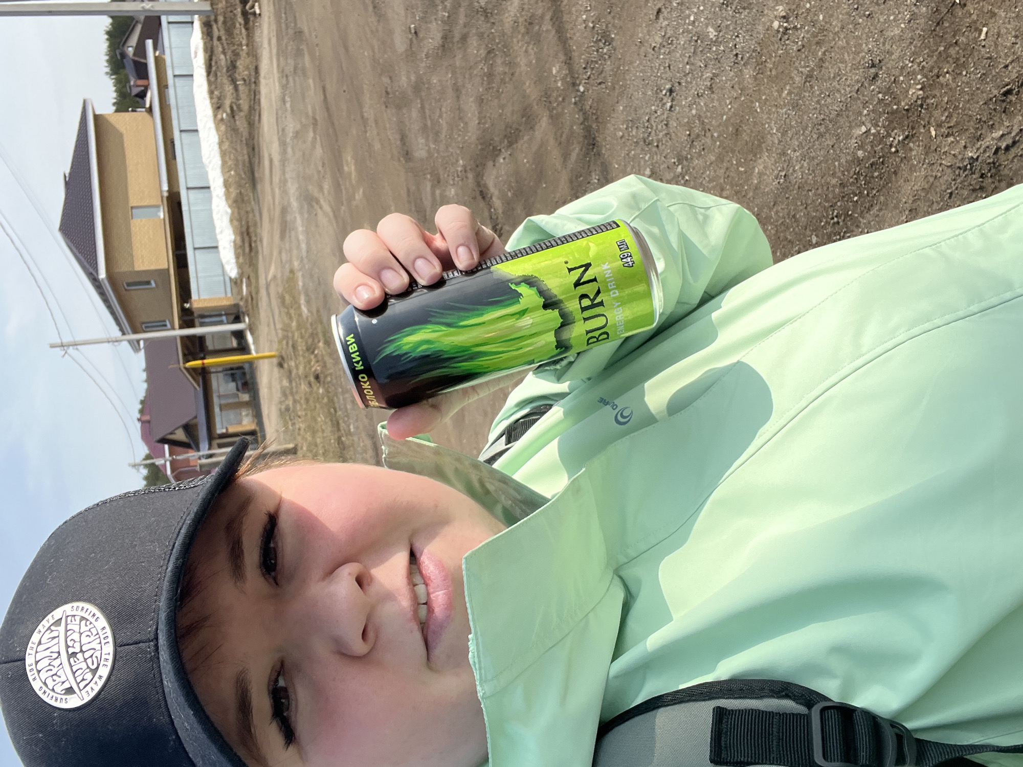 A person is holding a "Burn" energy drink in their hand, posing for a photo outdoors. They are wearing a black baseball cap with a white patch, and a light green jacket. The background shows a building and some dirt ground. The brand of the energy drink and the outdoor setting suggest a scene in the city of Chaikovsky, Russia, where the "Burn" energy drink is available and people engage in outdoor activities.
