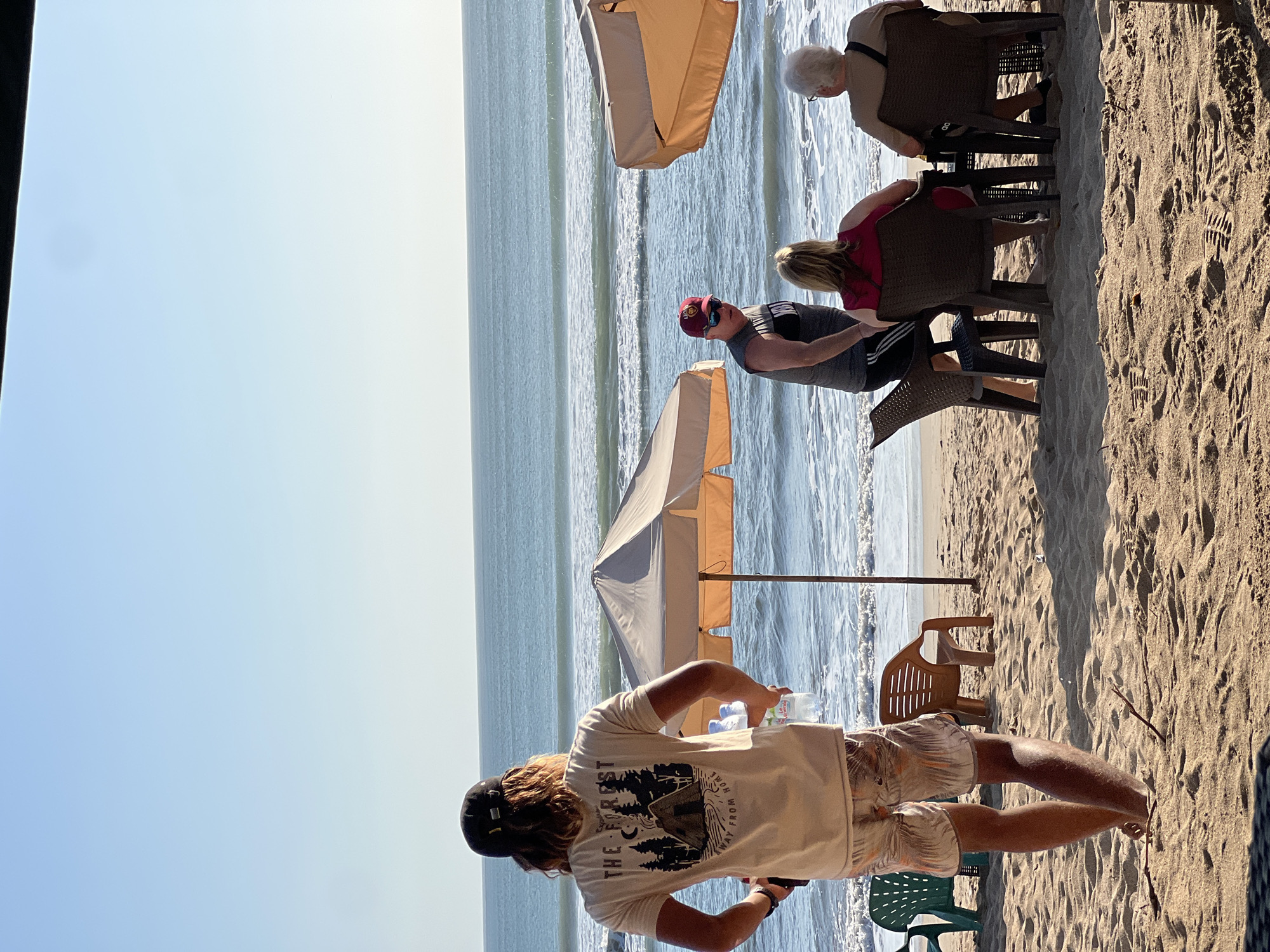 A man wearing a black baseball cap and a tan t-shirt with a graphic print stands facing the ocean on a sandy beach. He holds two bottles of water. In the background, waves crash on the shore. Several beach umbrellas and chairs are set up on the sand. To the right, a man in a gray tank top and red baseball cap is standing and talking to two people, a woman in a pink shirt and an older person in a tan shirt, who are seated at a table. The scene appears to be a beach cafe or restaurant in Kuta, Indonesia, given the ocean and beach setting.
