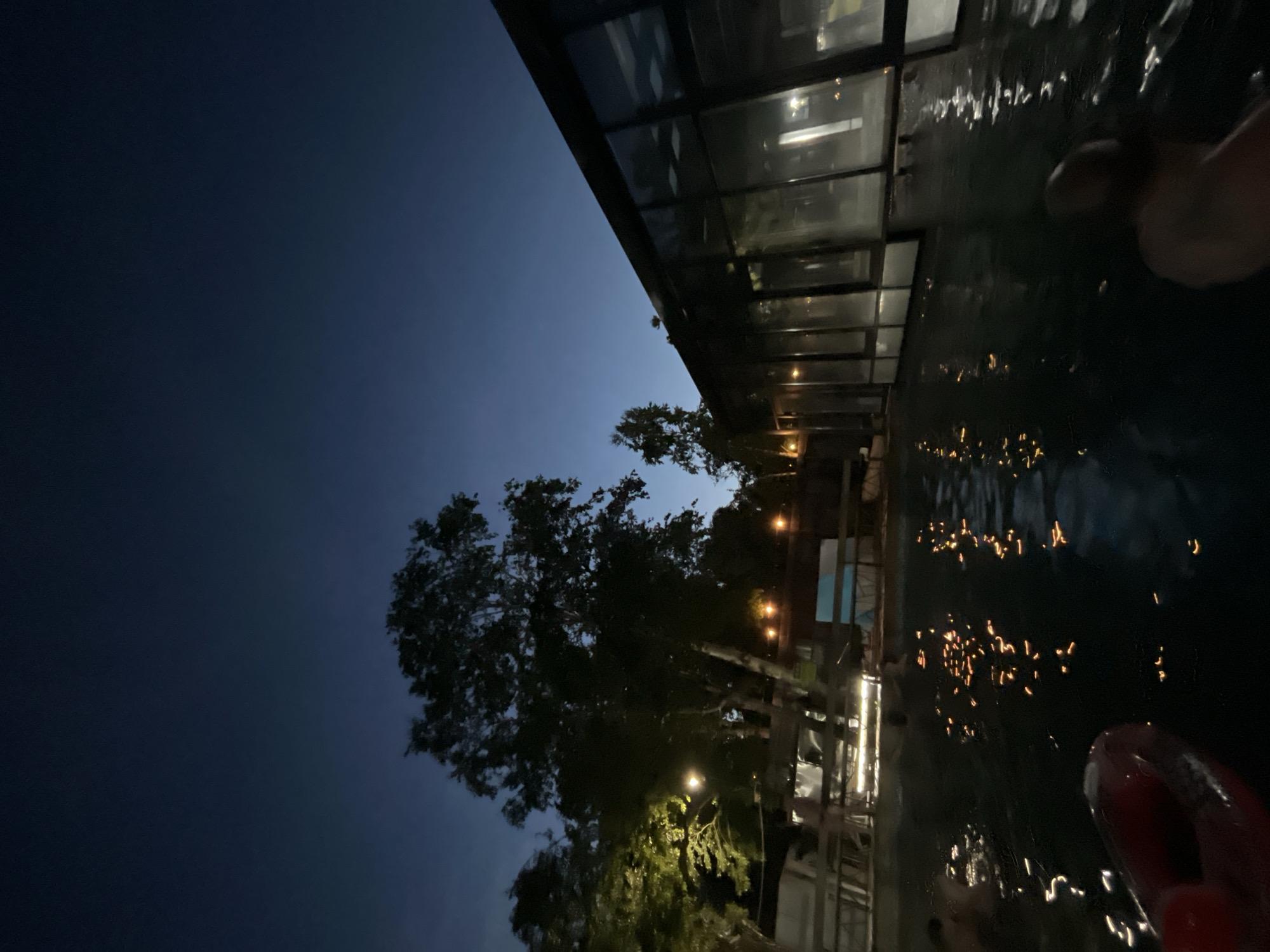 The image depicts an outdoor swimming pool at night in Paratunka, Russia. The sky is a deep twilight blue, with a gradient towards black at the top of the frame. Silhouetted against the sky are several trees, one with distinctive white bark, possibly birch. To the right, a modern building with large, dark windows stretches across the frame, reflecting some of the interior lights and the twilight sky.
The pool itself is dark, with the water surface reflecting scattered orange lights from an unseen source, possibly submerged lights or lights from the surrounding area. There are faint ripples on the water's surface. In the foreground, a red inflatable ring or buoy is partially visible in the lower left corner. In the water, several people are present, though mostly obscured by the darkness and the water. One person's shoulder and head are visible in the lower right, and others are seen further back in the pool, appearing to swim or relax. Some structures, possibly walkways or changing rooms, are visible above and behind the pool area, with lights illuminating parts of them. The overall atmosphere is serene and cool, characteristic of an evening swim.