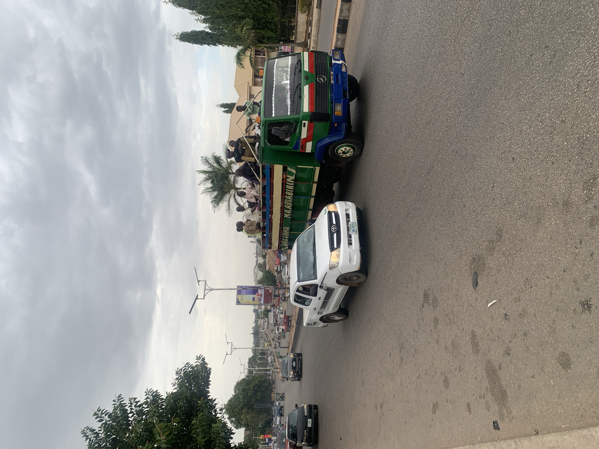 This image captures a busy street scene in Jos, Nigeria, under an overcast sky. The central focus is a large, older model Mercedes-Benz truck, distinctively painted green with a blue front bumper and cabin, accented with red and white stripes. Numerous people, primarily men, are seen riding in the open bed of this truck, standing and sitting. Some individuals are also visible near the truck's cabin. The text "M. A. ASSABIRIN" is visible in green on the side of the truck bed.
Adjacent to the green truck, a white Toyota pickup truck is traveling, its license plate "YNN 70 CAA" partially visible. Further down the asphalt road, a line of other vehicles, including cars and vans, suggests ongoing traffic.
The background shows urban and natural elements: buildings, streetlights, and various trees, including palm trees. The sky is filled with light grey clouds, indicating an overcast day without direct sunlight. The scene portrays typical daily transport and activity in a bustling environment.