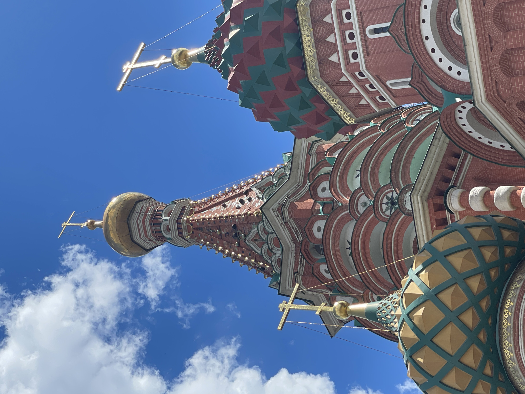 The image shows a low-angle shot of the ornate, multi-domed St. Basil's Cathedral in Moscow, Russia. The sky is a vibrant, clear blue with scattered white clouds, suggesting a bright and sunny day. The tops of several onion domes, adorned with intricate patterns of red, green, and gold, are visible. Golden crosses surmount some of the domes. The architectural style is distinctly Russian Orthodox, characterized by its colorful, patterned domes and rich detailing in brick and decorative elements. The scene captures the grandeur of the cathedral, with the bright sunlight highlighting its vibrant colors and complex textures. The perspective emphasizes the height and majesty of the structure against the expansive sky. There is no visible text.