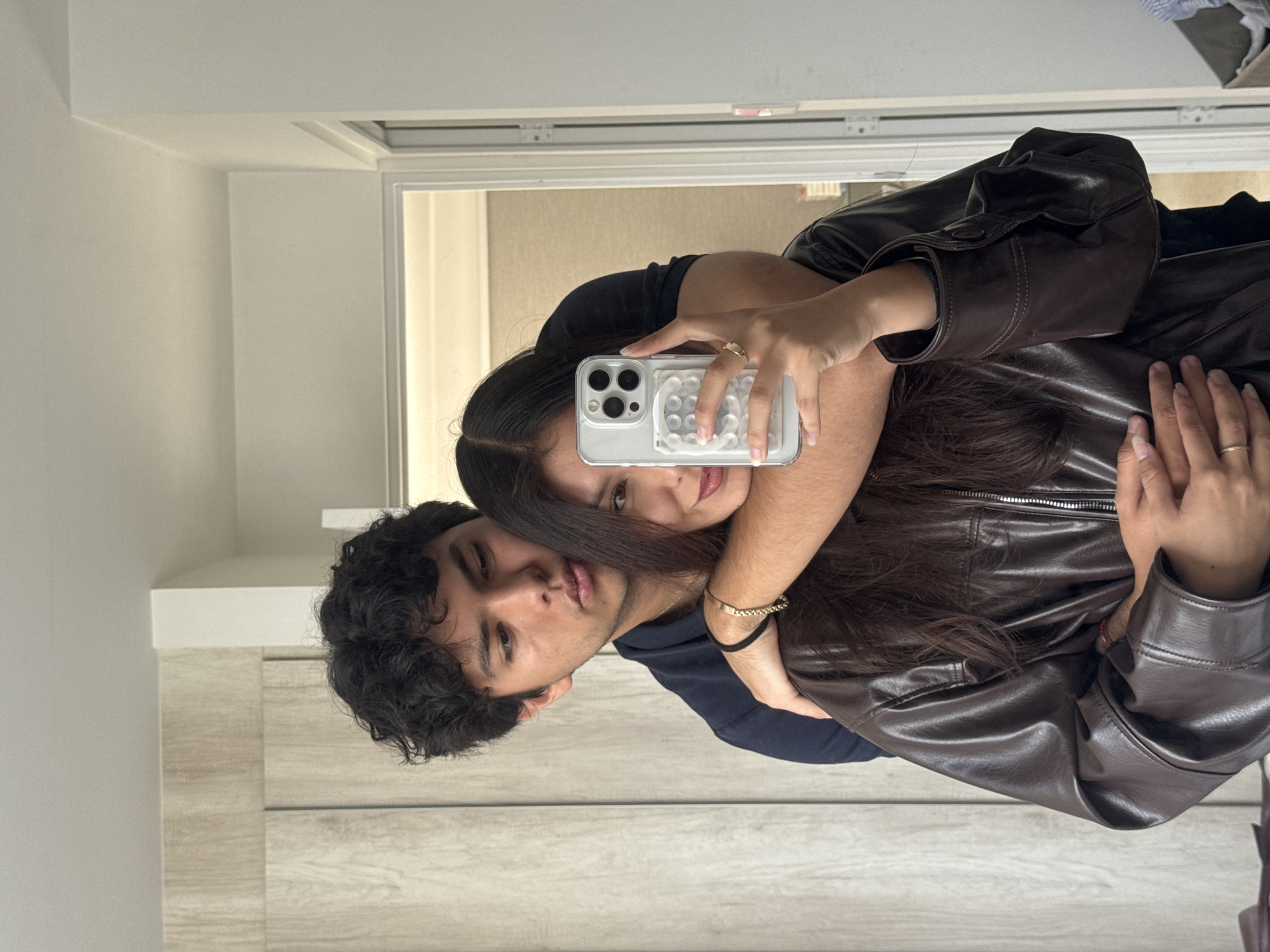 A young couple poses for a selfie in what appears to be a bathroom or bedroom. The man has dark curly hair and is wearing a navy blue shirt. The woman, with long dark hair, is embracing him from behind while holding a smartphone up to capture the image. She is wearing a brown leather jacket. The phone has a clear case with a white, bubble-like design. There is a gold ring on her finger. The scene is indoors, with a door and light-colored furniture in the background. The photograph appears to have been taken in portrait mode.