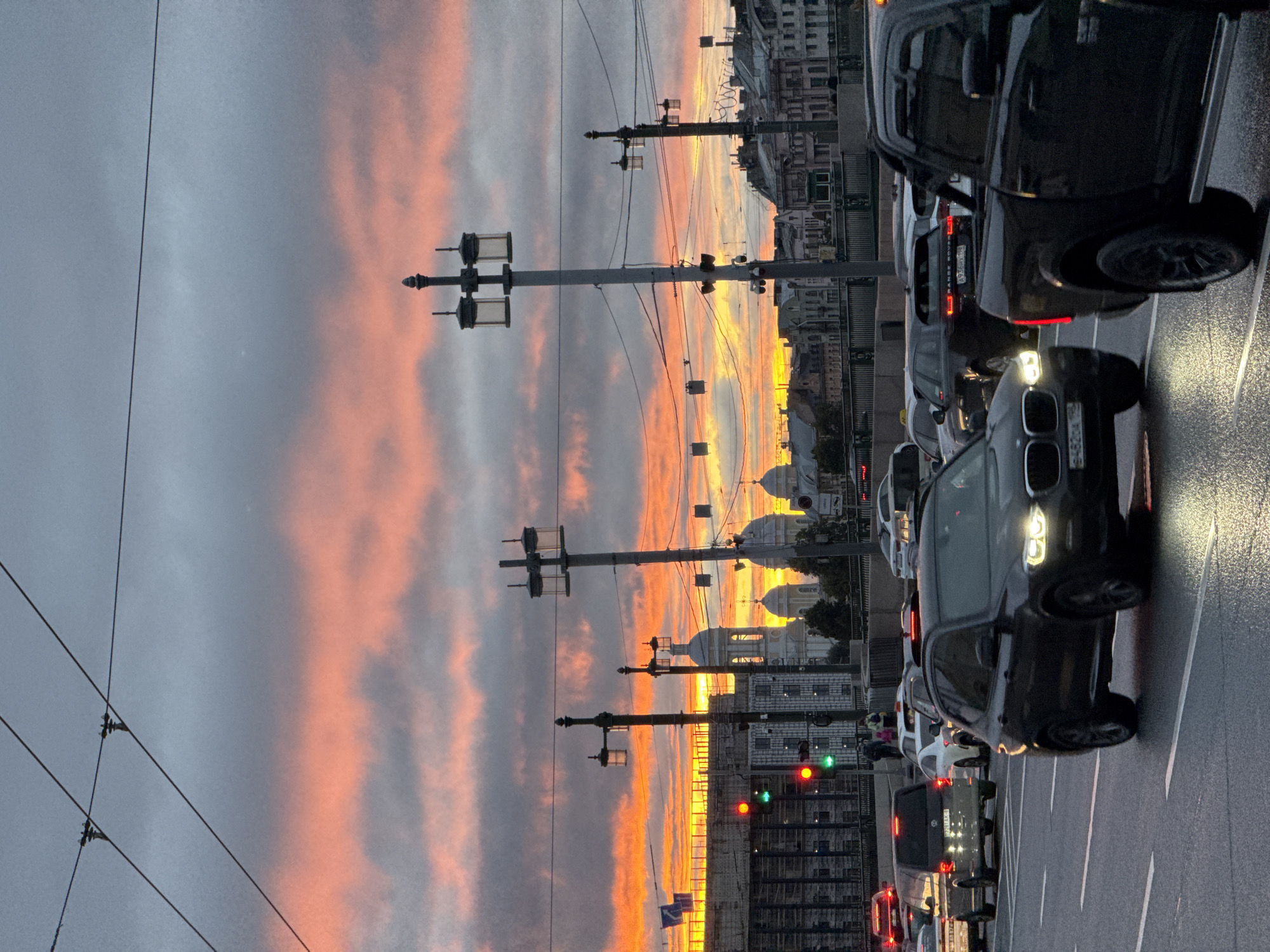 This media file depicts a street scene in Saint Petersburg, Russia, during what appears to be sunset. The sky is filled with dramatic clouds tinged with orange and pink hues, contrasting with the gray clouds. Several tall lampposts with multiple lights are visible, running along the street. In the foreground and midground, there is a line of cars, suggesting traffic congestion. One car, a black SUV, is particularly prominent, with its headlights illuminated. The road appears wet, possibly indicating recent rain. In the background, the architecture of Saint Petersburg can be seen, including buildings with domed roofs, characteristic of the city's historical structures. Traffic lights are also visible, with red and green lights illuminated. The overall mood is somewhat somber due to the overcast sky and traffic, but the vibrant sunset adds a touch of beauty and drama to the scene. No visible text is clearly legible.